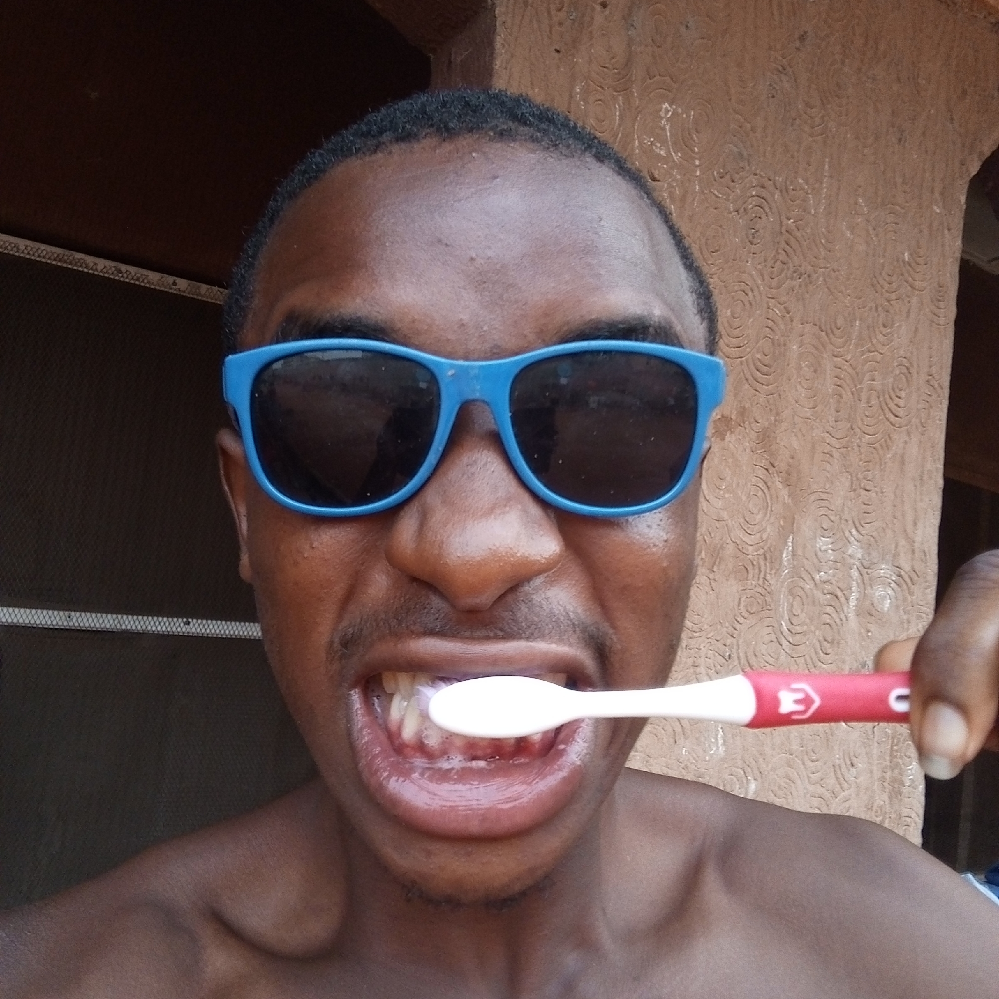 An African male with short hair and wearing blue sunglasses is captured at close range brushing his teeth with a red and white toothbrush. The background reveals a textured brown wall to his right and a window or opening with a screen to his left. It is not possible to determine the precise location within Onitsha, Nigeria from this image alone.