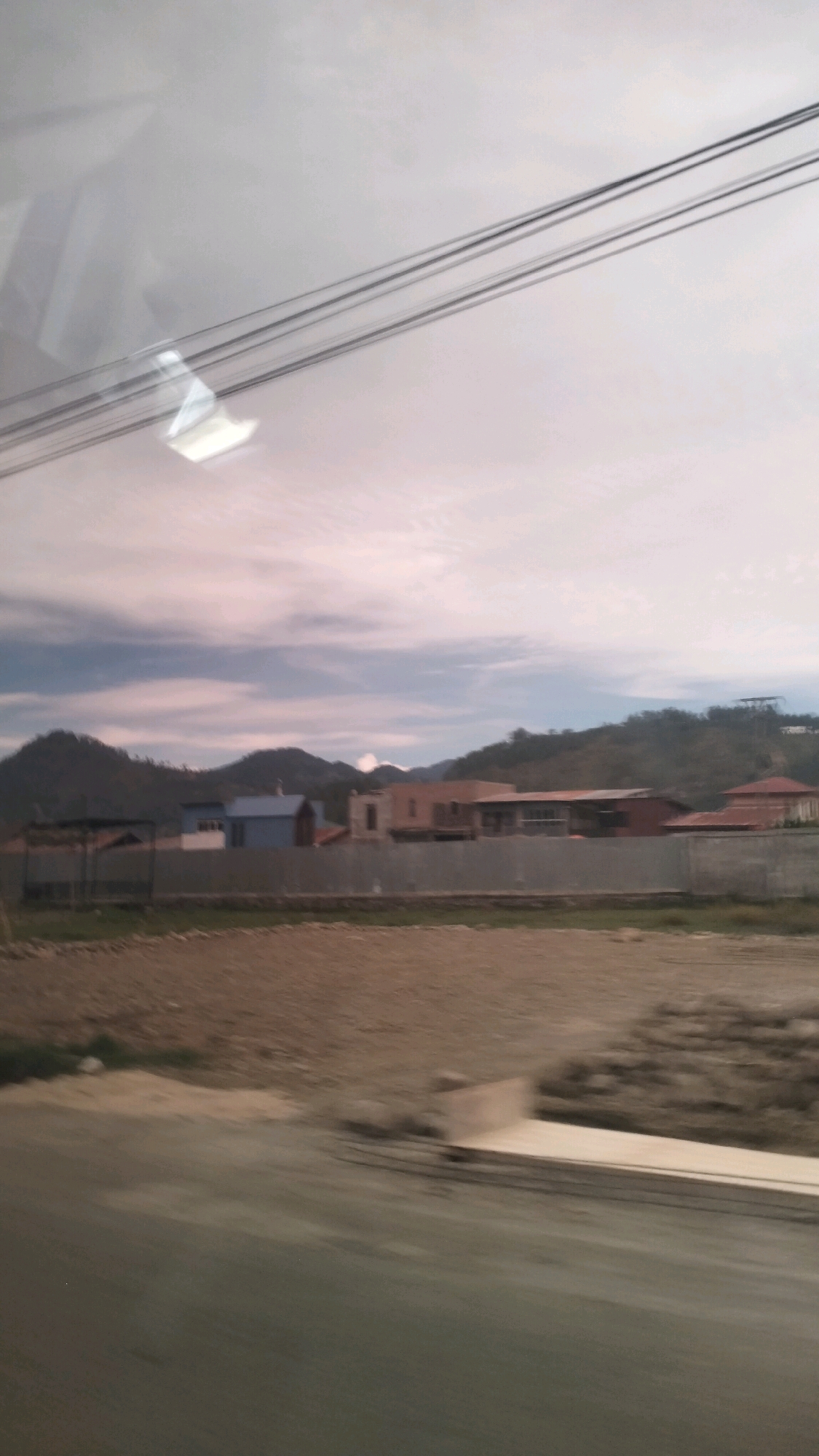 The image depicts a blurry, daytime outdoor scene, likely taken from a moving vehicle, suggesting a roadside view. The foreground is dominated by a blurred patch of ground, possibly a road or dirt path, with indistinct shapes of what could be construction materials or debris. Further back, a strip of brown earth stretches across the frame, bordered by a low, grey fence. Beyond the fence, a row of buildings is visible, varying in color and architectural style, with some featuring terracotta-colored roofs. These buildings appear to be residential or commercial structures. The middle ground is characterized by a landscape of rolling hills and mountains, some of which are covered in dense vegetation. The sky is overcast with wispy clouds, and the overall lighting suggests either early morning or late afternoon. A prominent element in the upper portion of the image is a set of parallel wires, likely power lines, running diagonally across the frame. The image's low resolution and blurriness make specific identification of people or precise actions difficult. The scene, with its mix of built structures and natural landscape, is consistent with a view from a road on the outskirts of a town or city, potentially in a semi-rural or developing area. While the request specifies the city of Kalakemili, Indonesia, there are no visually identifiable landmarks or specific details within the image to confirm this location.