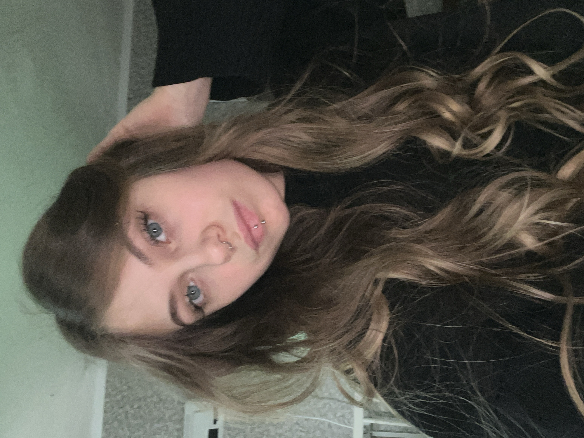 The image is a vertically oriented photograph of a young woman. She has long, wavy, light brown hair with blonde highlights. Her eyes are light blue. She has piercings in her left nostril and lower lip. She is wearing a black top. Her left hand is resting on her head. The background is out of focus but shows a pale green wall and a patterned floor. No evidence in the image suggests it was taken in the city of Фоки, Russia.