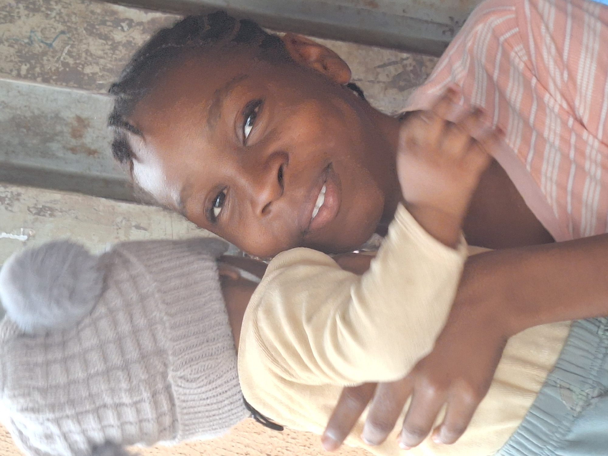 A medium shot, indoors. A Black woman lies sideways, cradling an infant. The woman is looking at the camera with a slight smile. She is wearing a striped shirt. The infant is facing away from the camera and wearing a light-colored outfit and a hat with a pom-pom. The background appears to be a rough, unfinished surface. The location is Sabon Tasha, Nigeria.
