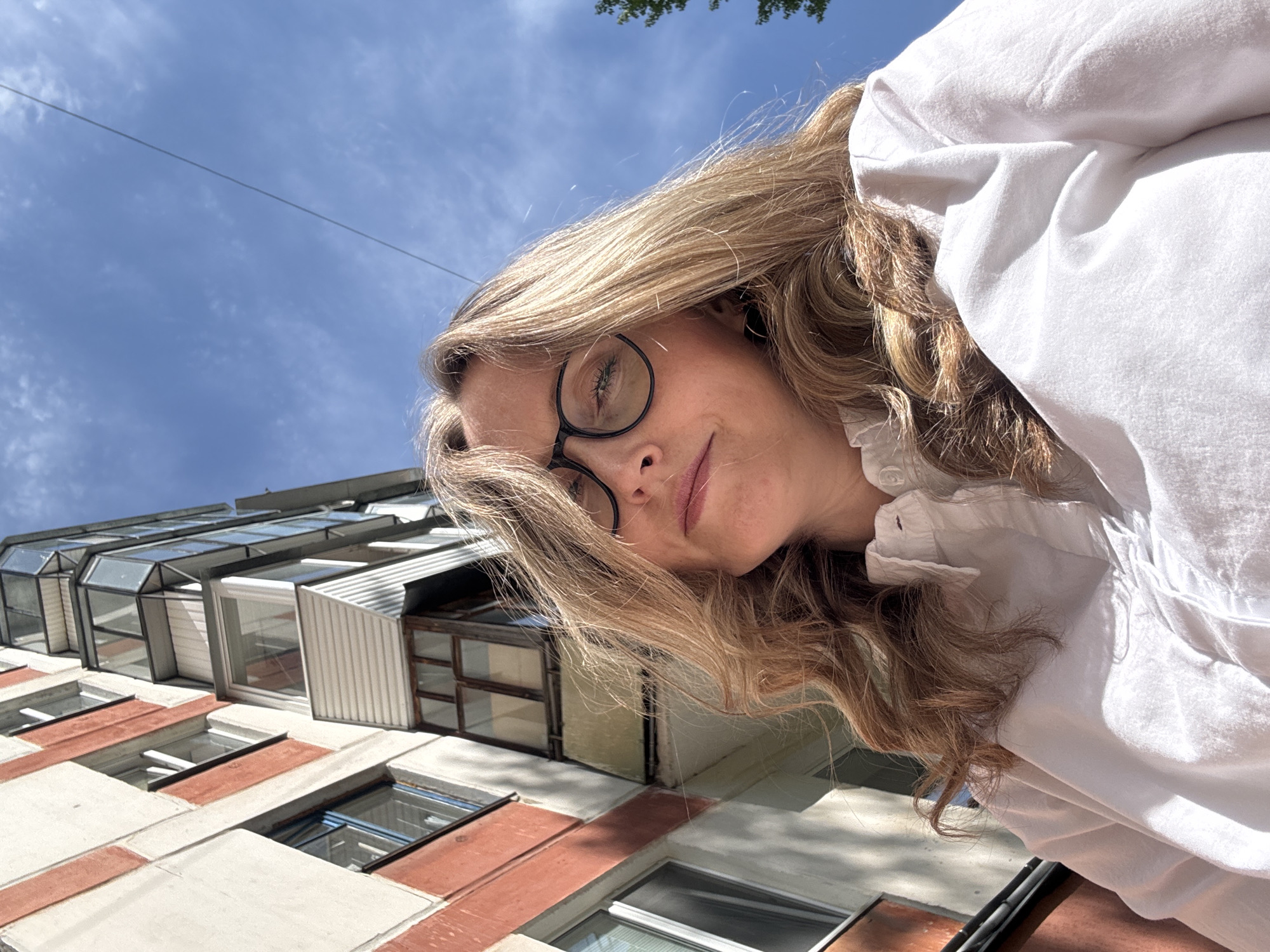 A woman with blonde, wavy hair and black-rimmed glasses is centered in the image, looking down at the camera. She is wearing a white blouse. Behind her, a light-colored building with red brick accents is visible, as well as a blue sky with light clouds. Tree leaves are visible at the top of the image. Based on the building style, this is likely in an urban area of Chaikovsky, Russia.