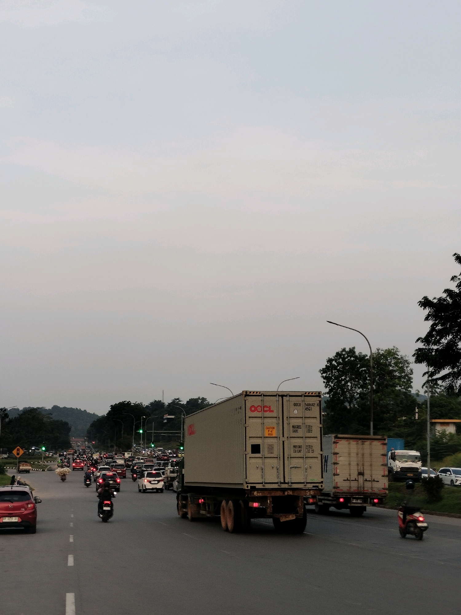 The image depicts a busy highway in Batam, Indonesia, likely during late afternoon or early evening, indicated by the overcast sky and traffic congestion. The foreground shows a two-lane asphalt road with white dashed lines. Several vehicles are visible, including a red hatchback on the left, a motorcycle in the center lane with a rider, and two large container trucks in the right lanes. The truck on the right is a flatbed trailer, while the one in the center has a large container unit labeled "OOCL" and another with "IW." In the background, there is a significant amount of traffic, including cars, motorcycles, and other trucks, suggesting a rush hour or heavy traffic situation. Traffic lights are visible in the distance, appearing to be red, contributing to the standstill. Lush green trees and hills form the backdrop, with some buildings and infrastructure like streetlights also present. The overall scene captures a typical urban traffic environment in Batam.