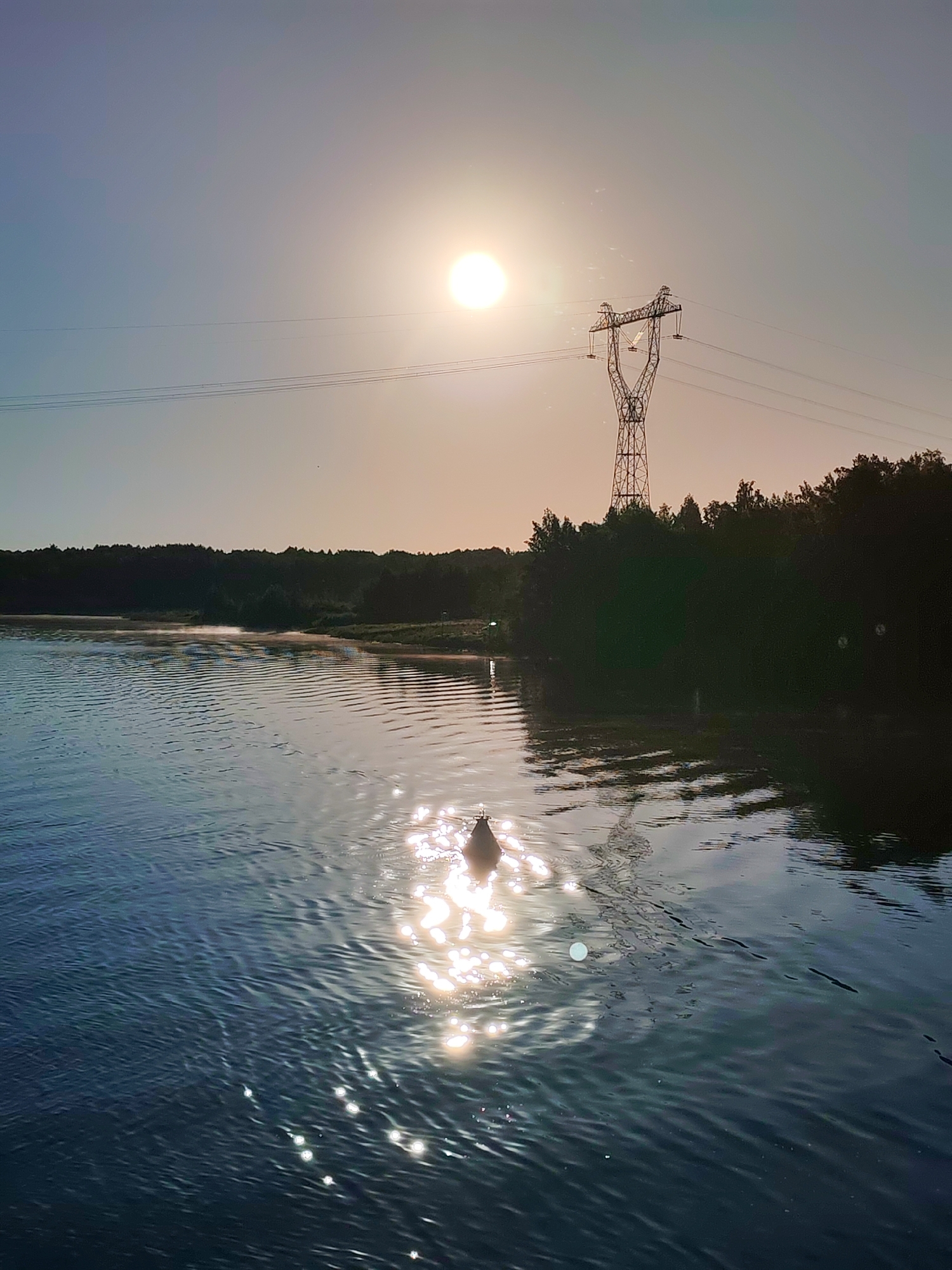 The image displays an outdoor scene in the city of Gari, Russia, characterized by strong sunlight. A large body of dark blue-grey water occupies the foreground, exhibiting ripples across its surface. A dark, bell-shaped buoy floats in the lower-middle, positioned within the path of intense sun glare reflecting off the water. The sun, appearing as a bright, circular disc, is situated in the upper center-right portion of the sky, which transitions from a light yellow-orange near the horizon to a pale blue-grey overhead. A dense, dark treeline forms the distant bank. A metallic electricity transmission tower stands prominently on the right, extending above the treeline, with multiple power lines visible stretching horizontally across the upper frame. No individuals or animals are present in the scene.