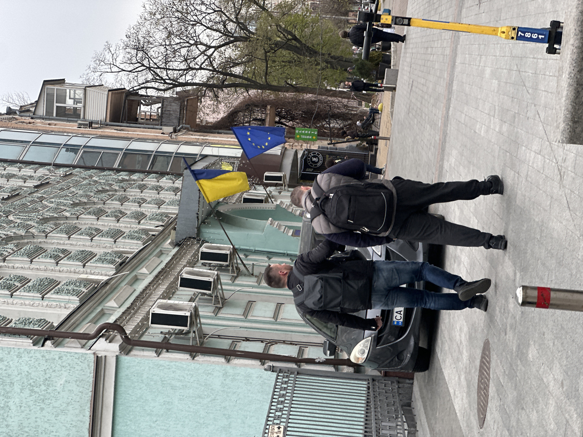 An urban street scene unfolds on an overcast day, likely springtime given the bare and budding trees. The perspective is from a slightly elevated position looking down a sidewalk lined with buildings.
In the foreground, two men are standing on the paved sidewalk. One man, seen from the side, wears a black jacket or vest over a long-sleeved shirt, blue jeans, and black shoes, standing next to a dark-colored parked car. Part of the car's Ukrainian license plate, showing "UA" below "... CA", is visible. Beside him, another man seen from behind wears a black jacket and a black backpack. Both men appear to be looking forward or slightly upwards. To the right, a silver bollard with a red top is visible.
The building on the left features a distinctive light blue/green facade with ornate decorative elements and multiple external air conditioning units. Above this, a modern glass-paneled roof leads to an upper structure with what appears to be a partially constructed or weathered rooftop addition, featuring wooden elements and windows. Attached to this building, a Ukrainian flag (yellow and blue) and a European Union flag (blue with yellow stars) fly from a pole. A green sign with white Cyrillic text reading "ГОТЕЛЬ" (Hotel) is also affixed to the building, below which is another dark sign with lighter text.
Further down the street, additional indistinct figures are visible, some standing near a yellow and blue bollard which has the number "1897" printed on it (when viewed upright). The ground is a patterned grey pavement. A partial view of a metal fence or gate in the lower left shows the Cyrillic word "РОЗПОЧИНАТИ" (Starting/Beginning). The overall atmosphere is that of a regular street day in Kyiv, Ukraine, suggested by the flags, license plate, and Cyrillic text.