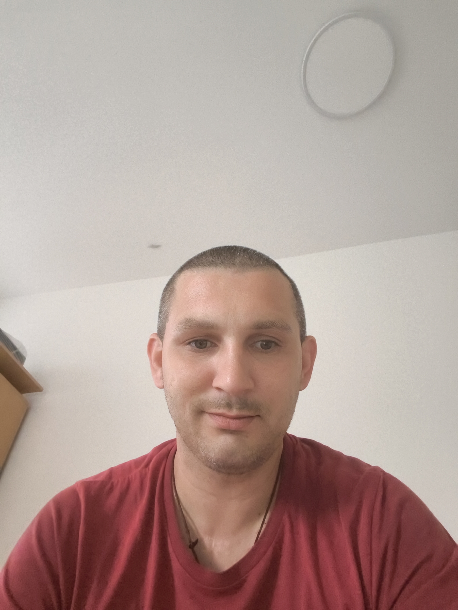 A close-up selfie of a man in his early 30s, with short brown hair and a light beard, wearing a red t-shirt. He is looking directly at the camera with a neutral expression. The background is a plain white wall and ceiling, with a circular light fixture visible on the ceiling. There is a hint of a wooden cabinet to the left of the frame. This image appears to be taken indoors, in a residential or office setting. There is no discernible information in the image that would indicate the location in the city of Wolgast, Germany.