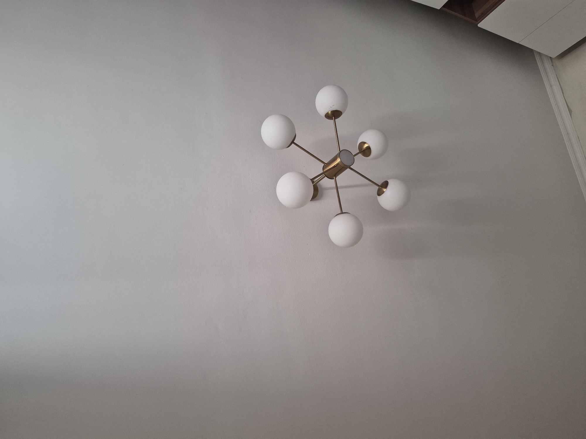 The image shows a close-up, upward view of a modern sputnik-style chandelier mounted on a plain, light-colored ceiling. The chandelier has a gold-toned metal frame with multiple arms extending outwards, each tipped with a white spherical glass globe. The globes are illuminated. To the right of the frame, a sliver of a white dresser with wooden accents is visible near the bottom edge. The lighting appears to be soft, creating subtle shadows on the ceiling. The scene is indoors, in what appears to be a residential or office space. There is no specific indication of the city of Bereke, Kazakhstan.