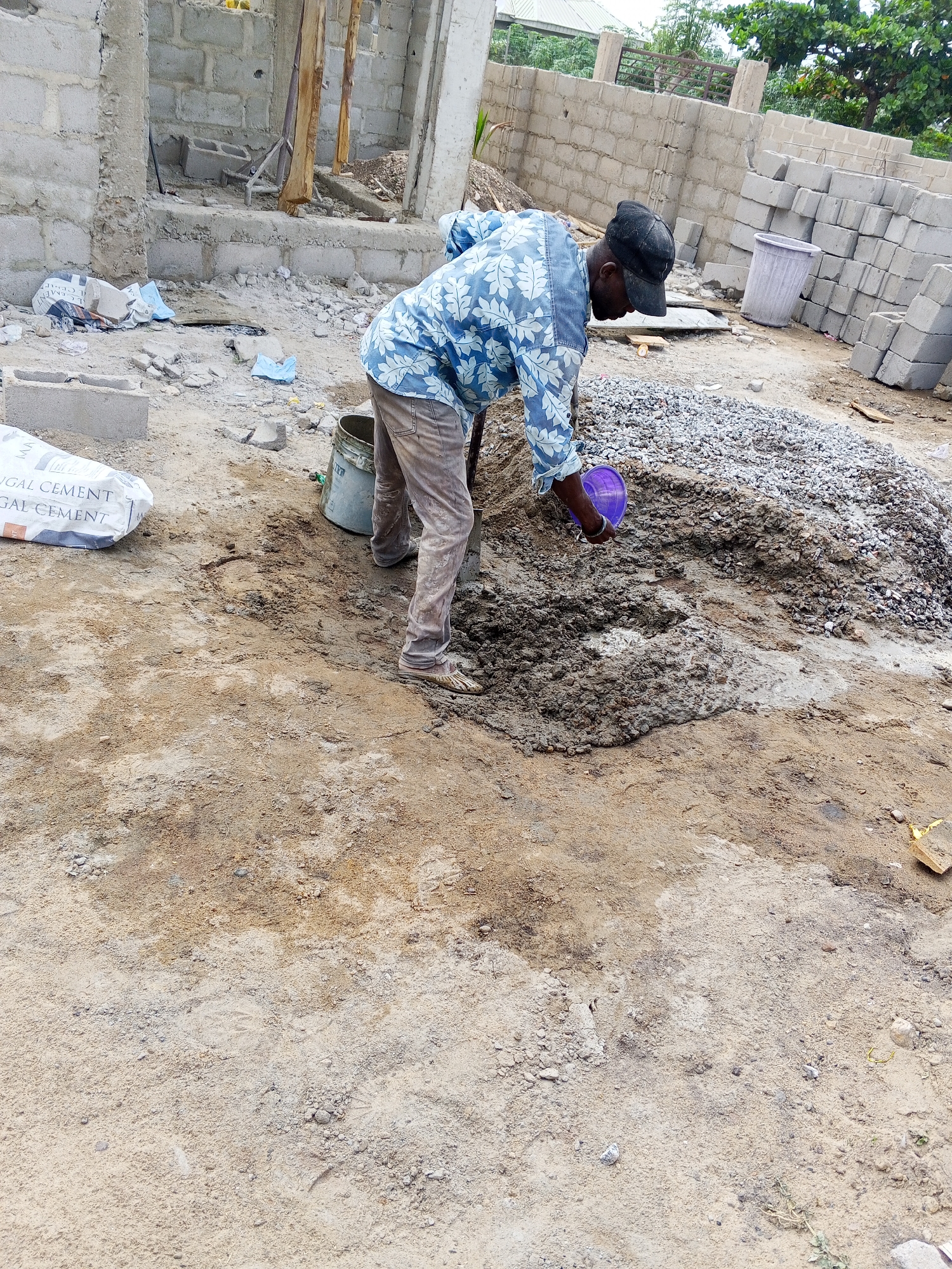 An outdoor scene captures a man engaged in manual labor at a construction site. The man, wearing a blue and white patterned long-sleeved shirt, light brown pants, and a black baseball cap, is bent over, mixing concrete or mortar on the ground. He is holding a purple plastic bowl or scoop, possibly adding water to the mixture, and a shovel handle is partially visible near his feet.
On the sandy, dirt ground, there is a large pile of gravel and sand, alongside a section of wet, grey concrete mixture that the man is working on. A grey bucket sits near his feet, and a white bag, partially visible on the left, bears the text "GAL CEMENT" and "GAT CEMENT," likely indicating a brand of cement.
The background shows the initial stages of building construction. Unfinished concrete block walls rise in various sections, with some wooden supports also visible. To the right, a large stack of grey concrete blocks is neatly piled, and a light purple or grey plastic bin stands nearby. Further in the distance, green foliage from trees suggests an outdoor, possibly rural or semi-urban, setting. The lighting is bright and even, indicating daytime, possibly under an overcast sky or during midday. The overall scene depicts a diligent worker focused on the task of preparing building materials.