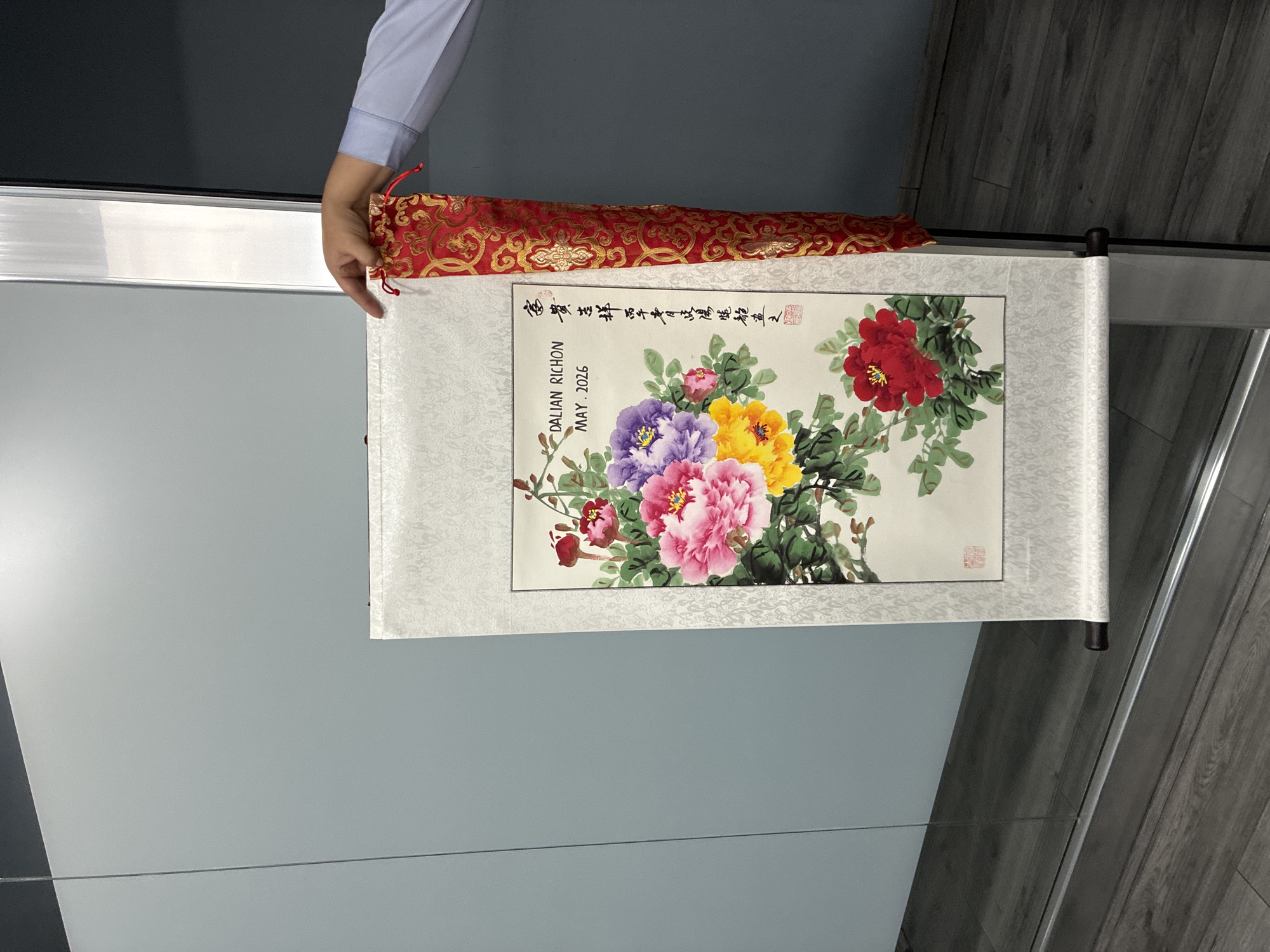 A person's hand is holding a red and gold brocade storage bag, above a partially unrolled scroll depicting a painting of flowers. The scroll is mounted on a patterned white border with wooden dowels at the top and bottom. The painting shows a bouquet of flowers, including red, pink, purple, and yellow blossoms, interspersed with green leaves. Calligraphic text in black ink and two red seals are visible at the top right of the painting. The words "DALIAN RICHON MAY. 2026" are printed in English on the left side of the text. The scene occurs indoors against a background of grey wall panels and grey wood flooring.