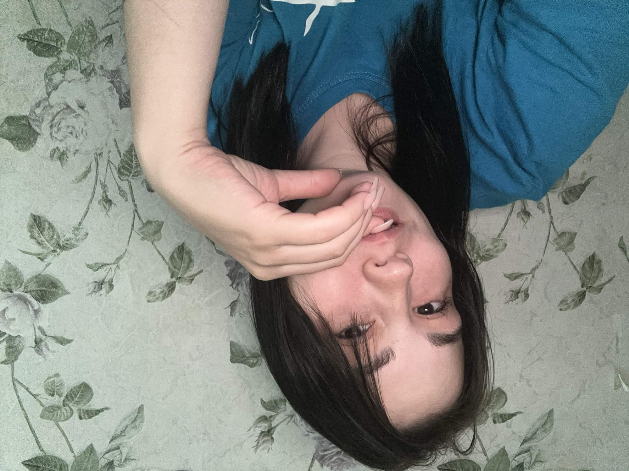 A woman with long, dark hair lies on her back against a light green floral patterned surface. She is wearing a teal shirt and has her left hand near her mouth, with her index finger touching her lips. Her face is angled towards the camera. The location is potentially in Chaikovsky, Russia, though this cannot be confirmed from the image alone.