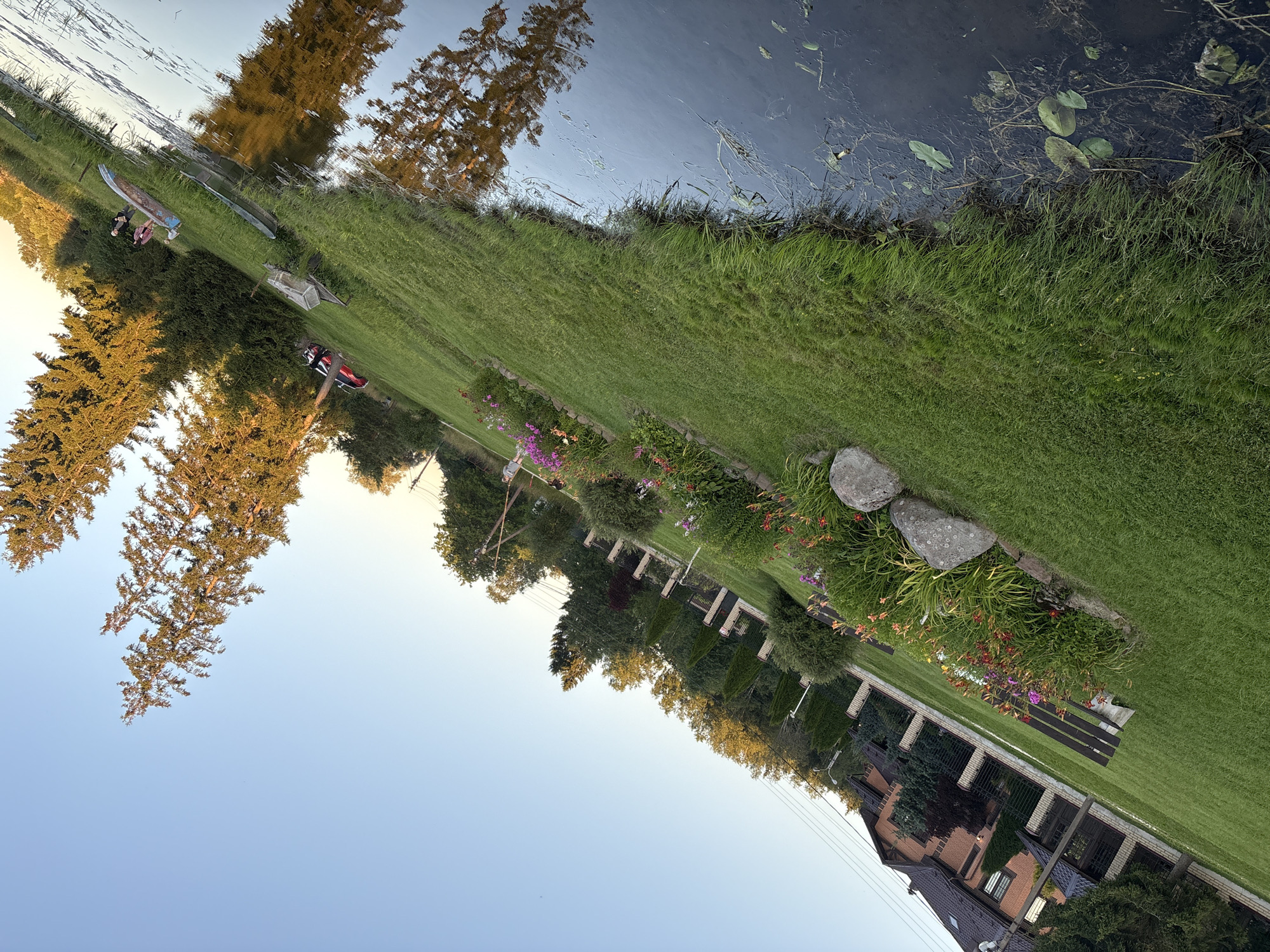 The image depicts a grassy yard adjacent to a dark body of water, likely a pond or slow-moving stream. Tall trees line the far bank of the water, their reflections shimmering on the surface. The yard itself is well-maintained, with neatly cut grass and a flower bed featuring various colorful blooms, including pinks and oranges. Two large rocks are situated near the flower bed. In the background, a red brick house with dark-tiled roofs and large windows is visible, bordered by a fence and more greenery. Several power lines traverse the sky. To the left of the water, a narrow boat is beached on the grass, and further down, a smaller boat or raft is also present. Two individuals are seated near the beached boat. The scene is bathed in the warm light of what appears to be late afternoon or early evening. The location is specified as Chikino, Russia.