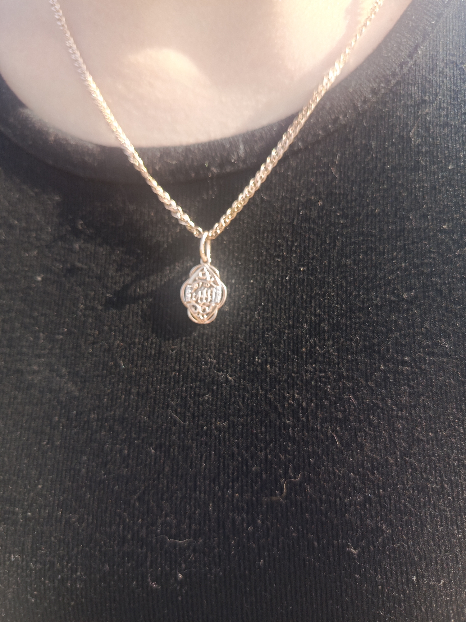 A close-up shot of a gold chain necklace with a clover-shaped pendant worn by a person. The chain is visible around the neck and extending down towards the center of the frame. The pendant hangs just below the collarbone. The person is wearing a black shirt or top. The photo is taken in natural light, with some areas slightly overexposed. The background is blurred and out of focus. Location: Unknown, Unknown.