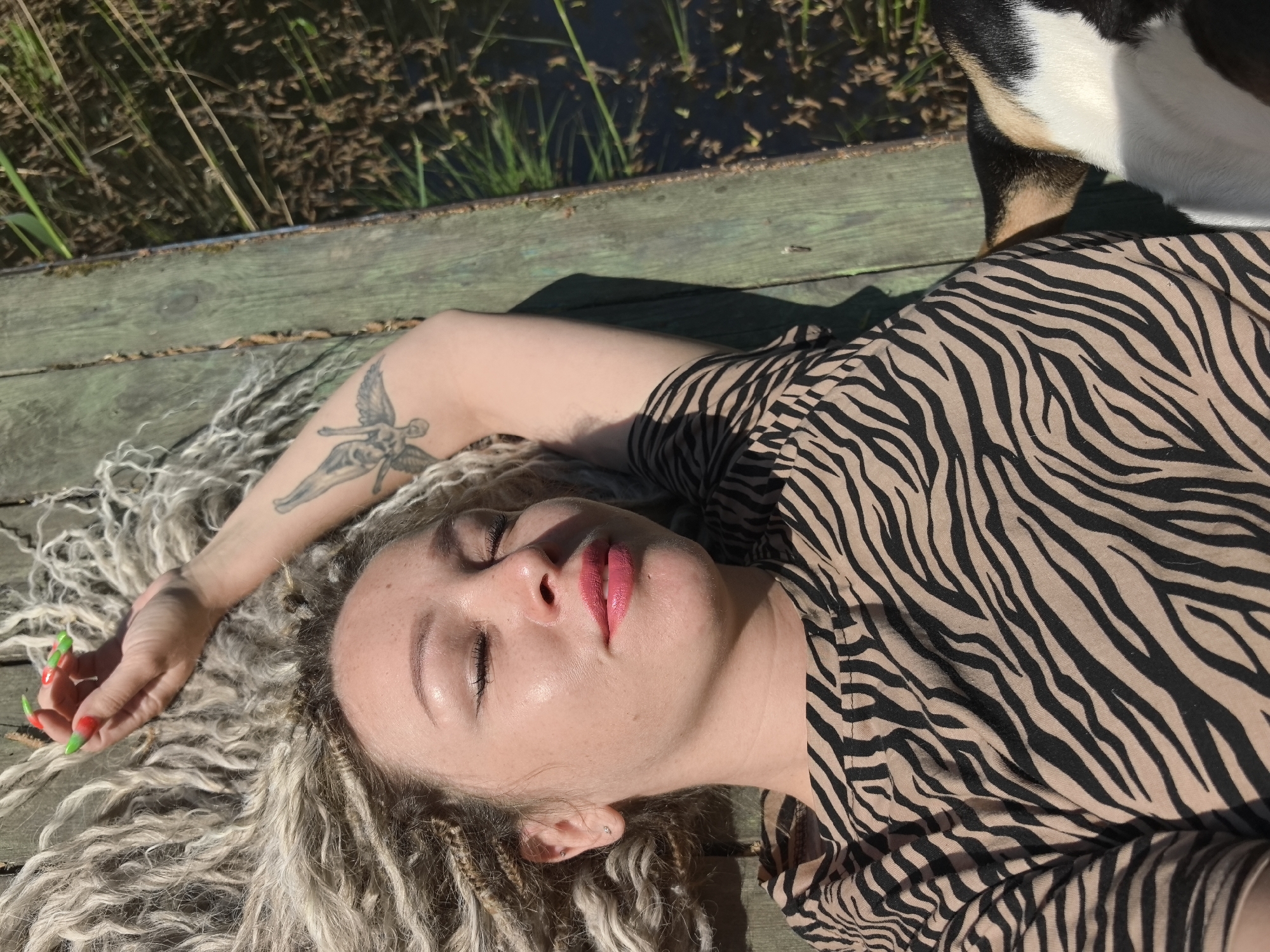 A woman is lying on a wooden pier beside a body of water. The pier appears to be aged and weathered. Patches of aquatic vegetation are visible in the water. The woman has light-colored, textured hair. She wears a zebra-striped top and her arm has a tattoo. The woman has red lipstick. A black-and-white animal is near her head, possibly a dog. Her nails are painted in different colors. The location is Noviy, Russia.