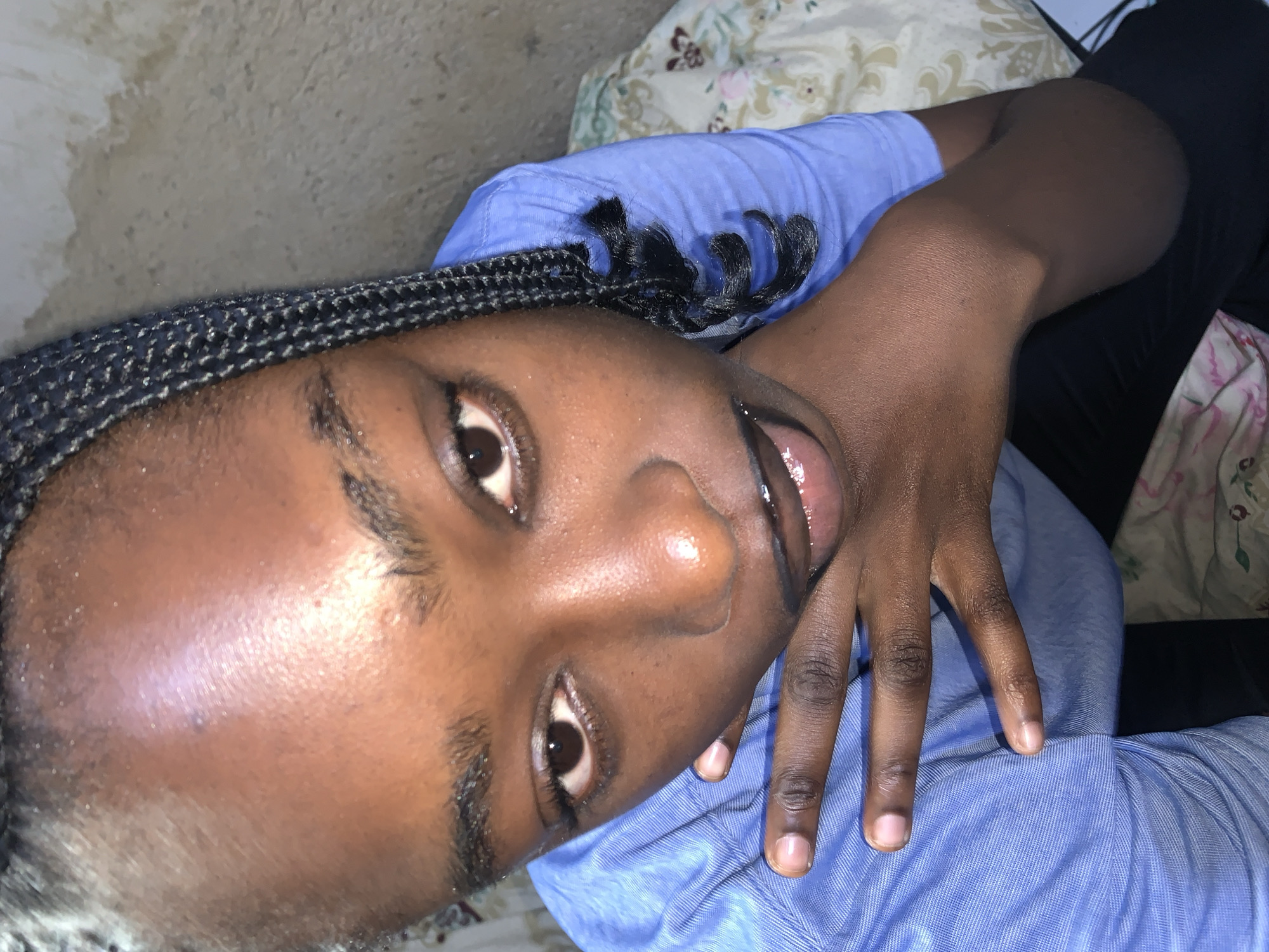 A close-up, overhead, medium shot shows a young Black person with braided hair, wearing a blue collared shirt, posing with their hand to their face. The person's left eye is visible, as is their nose and mouth, which is slightly open, revealing some teeth. The background consists of a textured wall and a floral patterned sheet. The scene is set in Rafin Par, Nigeria.