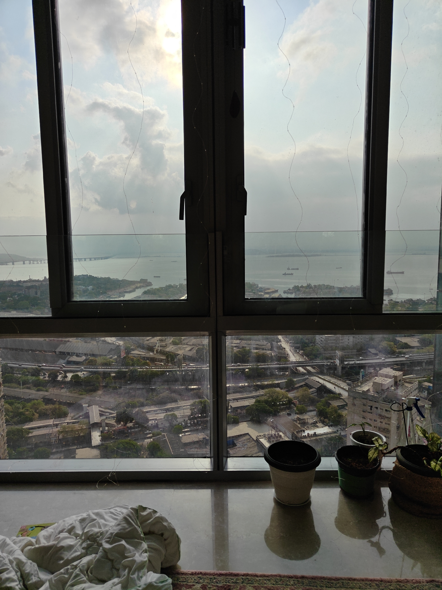 The image shows a view from a high-rise building in Mumbai, India, looking out over the city and the Arabian Sea. A large, multi-pane window dominates the foreground, offering two distinct visual layers: the upper panes frame a vista of the sea, with ships visible on the water, and the Mumbai Trans Harbour Sea Link stretching across the horizon. The lower panes reveal a dense urban landscape with roads, elevated highways, and buildings.
On the inside, three potted plants sit on the window sill. A rumpled white duvet or blanket is partially visible in the lower left corner, resting on a rug. The light outside suggests a cloudy or hazy day. The windows have thin, delicate lines that appear to be decorative.