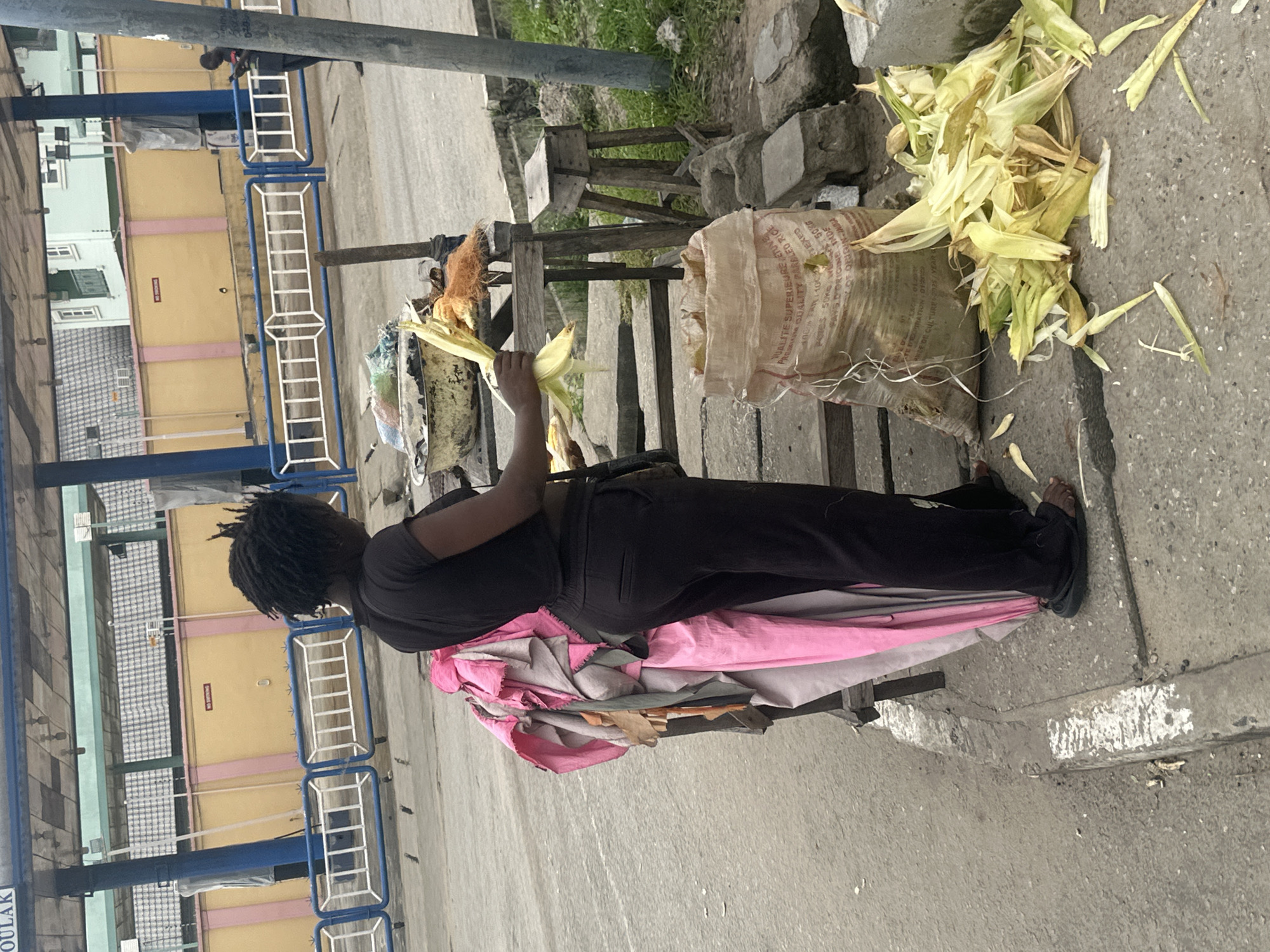 In Badagry, Nigeria, a woman with dreadlocks is standing next to a vendor's stall, holding several ears of corn. She is wearing a black t-shirt and black pants. The stall has a variety of items, including a large bag that appears to be filled with rice, and a pile of corn husks on the ground. In the background, there is a yellow wall with pink accents, and a blue barrier. Another person is visible in the distance, looking at their phone. The scene appears to be outdoors, possibly near a bus stop or a market.