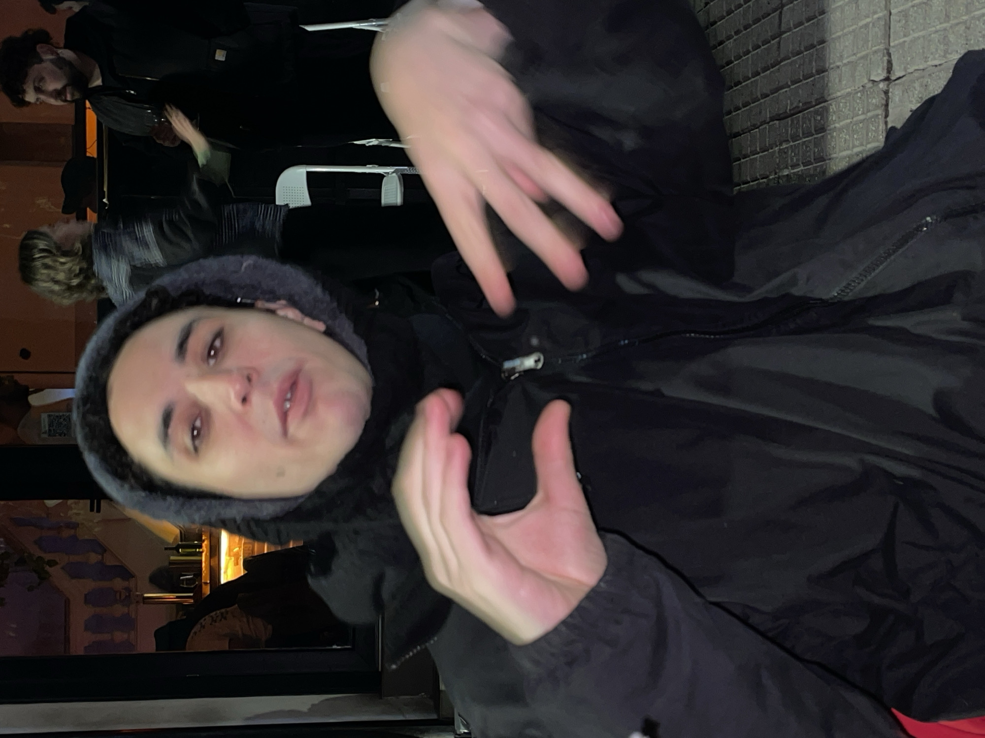 The image shows a person wearing a black hooded jacket, with their hands raised in a gesture. In the background, indistinct figures are visible at a building with lighted windows. The location appears to be in the city of Buenos Aires, Argentina.