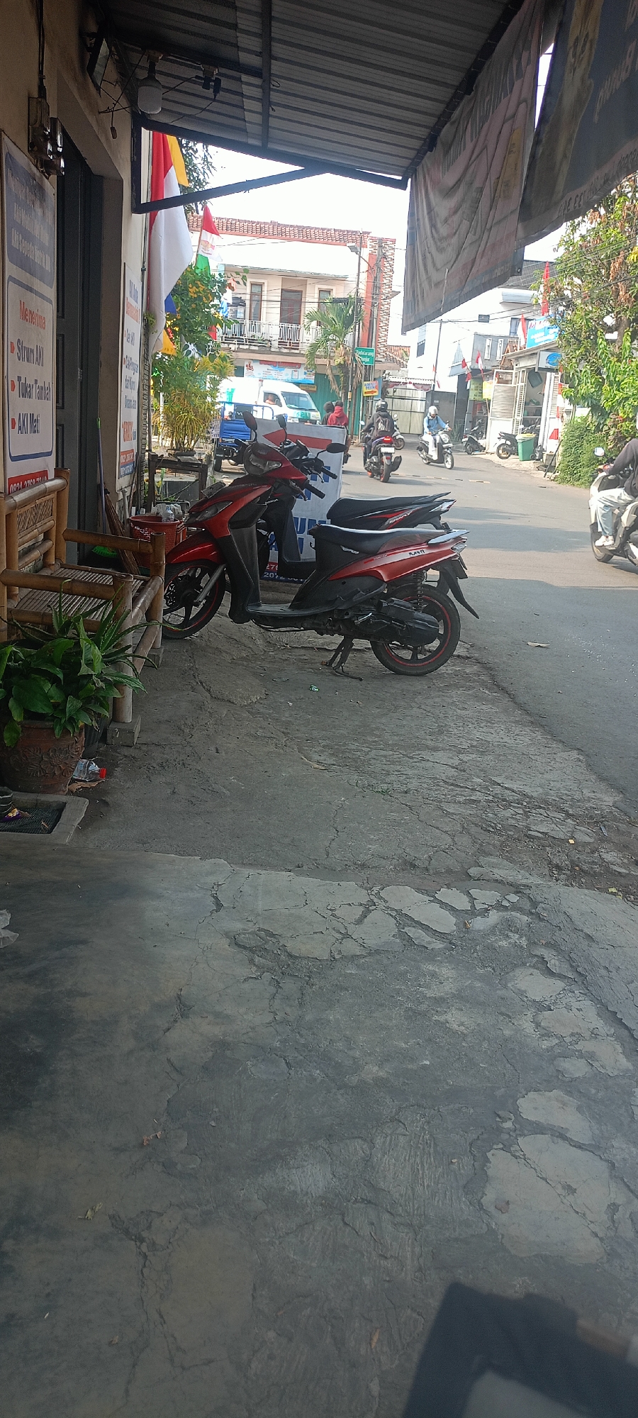 The image captures a street scene in Cimahi, Indonesia, likely in front of a business. A red and black scooter is parked on the side of the road. In the background, several other scooters and a white van are visible, suggesting a moderately busy street. A person is riding a scooter in the distance. There are also some buildings and trees lining the street, as well as flags of Indonesia and possibly other countries. A bamboo bench and some plants are in the foreground, next to a sign that lists services in Indonesian. The overall impression is of a typical day in a local Indonesian town.