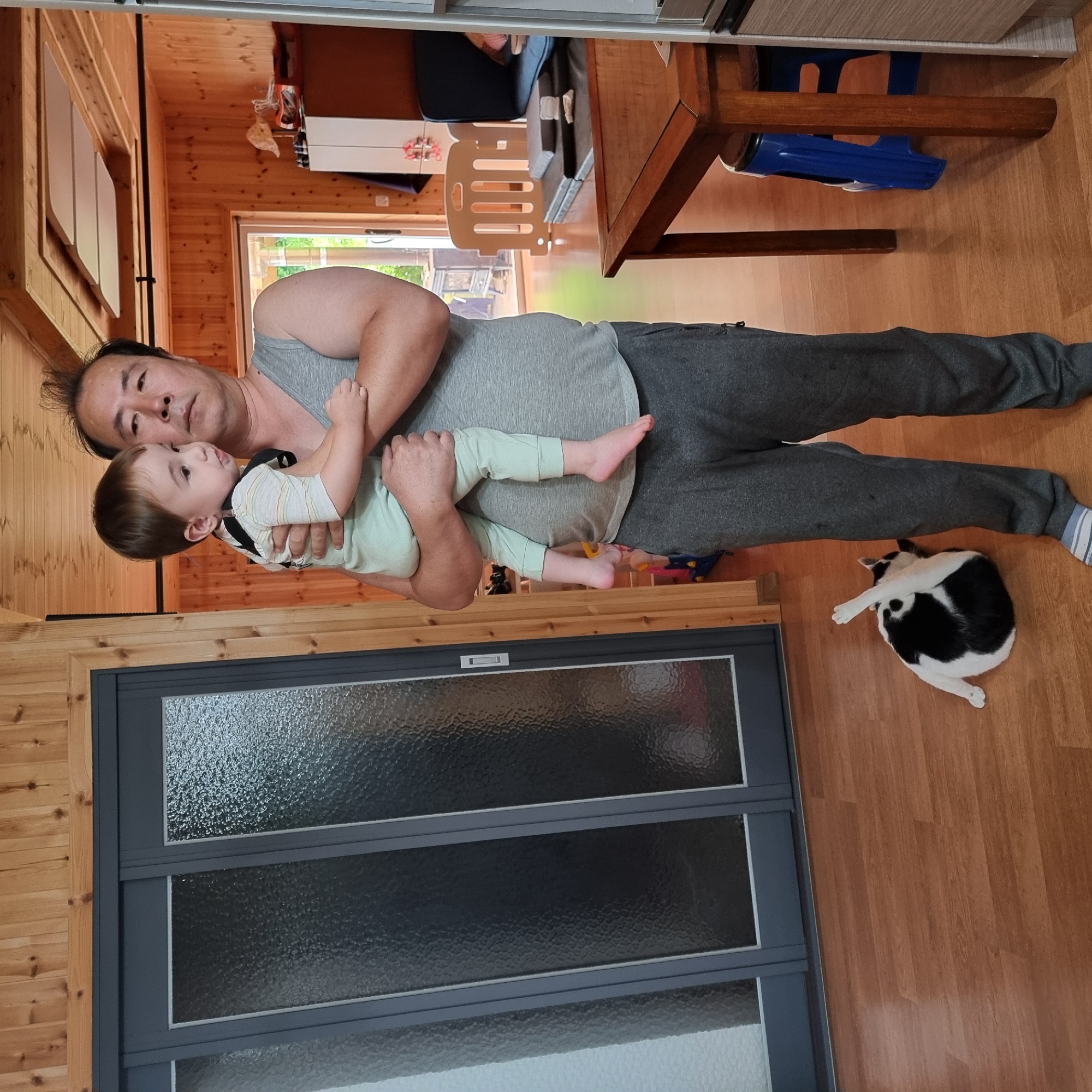 A man is holding a young child indoors. The man, wearing a gray tank top and gray sweatpants, is facing to the right and looking down at the child. The child, wearing a pale green outfit with stripes and a black bib, is being held in the man's arms and is also looking to the right. A black and white cat is lying on its back on the wooden floor, with its paws in the air, in the lower left portion of the image. The room has wooden paneling on the walls and ceiling. In the background, there are sliding doors with frosted glass, a wooden table with chairs, and a white cabinet. The image appears to be taken in a residential setting in Dunnae-myeon, South Korea.