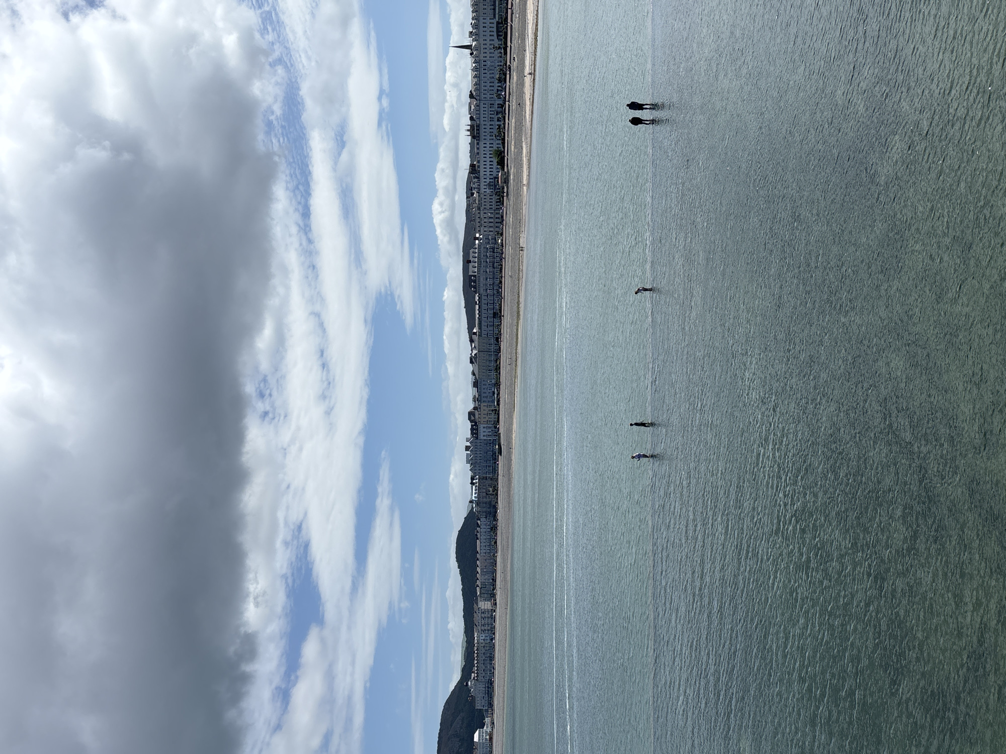The image depicts the beach and seafront of Llandudno, United Kingdom, under a sky of mixed blue and cloudy grey. A row of buildings lines the shore. Several figures stand in the sea at various distances from the beach. The sea appears relatively calm, with slight ripples on its surface.