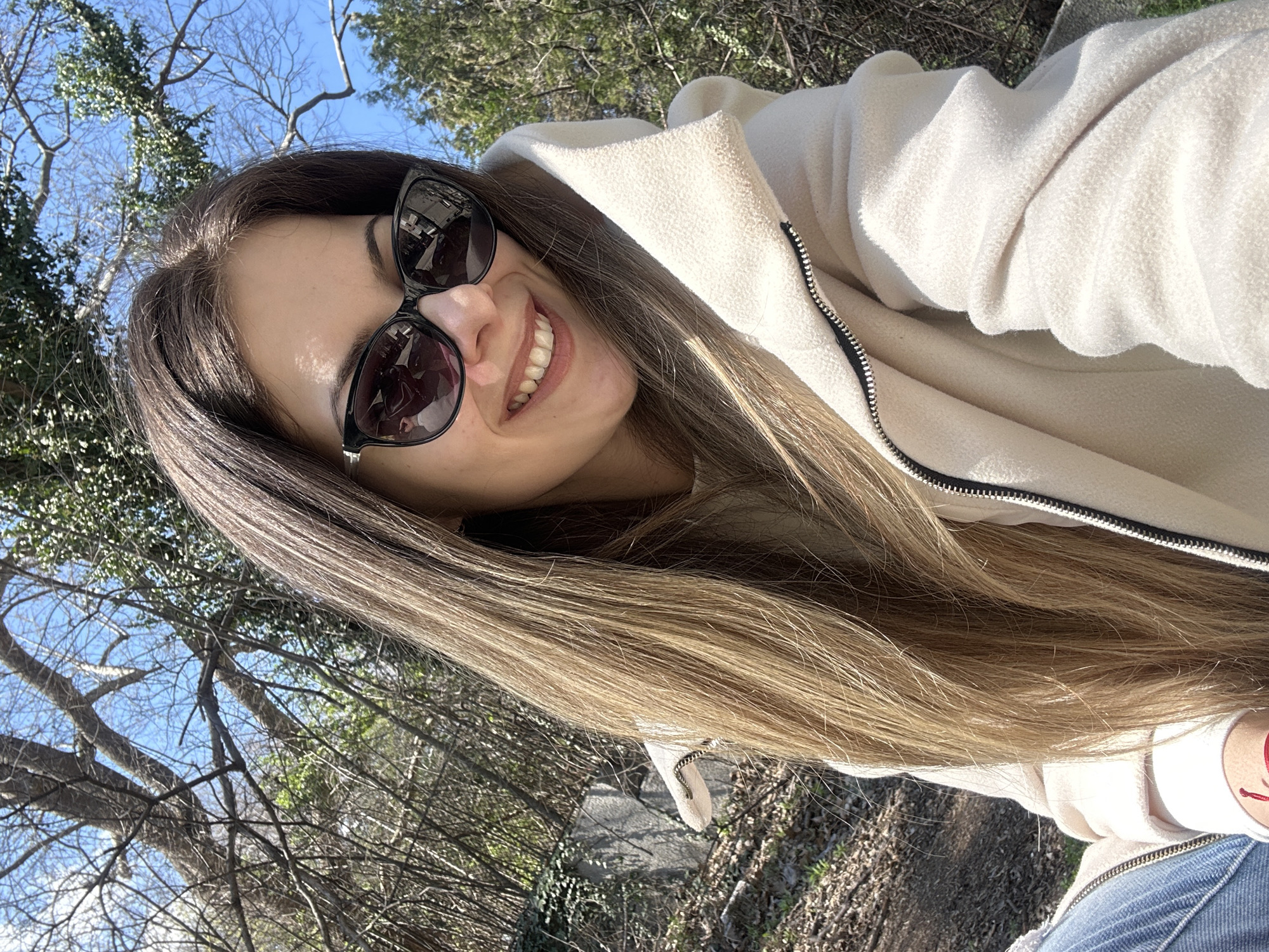 A woman is photographed from a slightly low angle in an outdoor setting. She is smiling, wearing sunglasses and a cream-colored hooded jacket. The scene includes trees and a blue sky, suggesting a park or wooded area. Without further details, it's impossible to pinpoint the exact location within Stara Zagora.