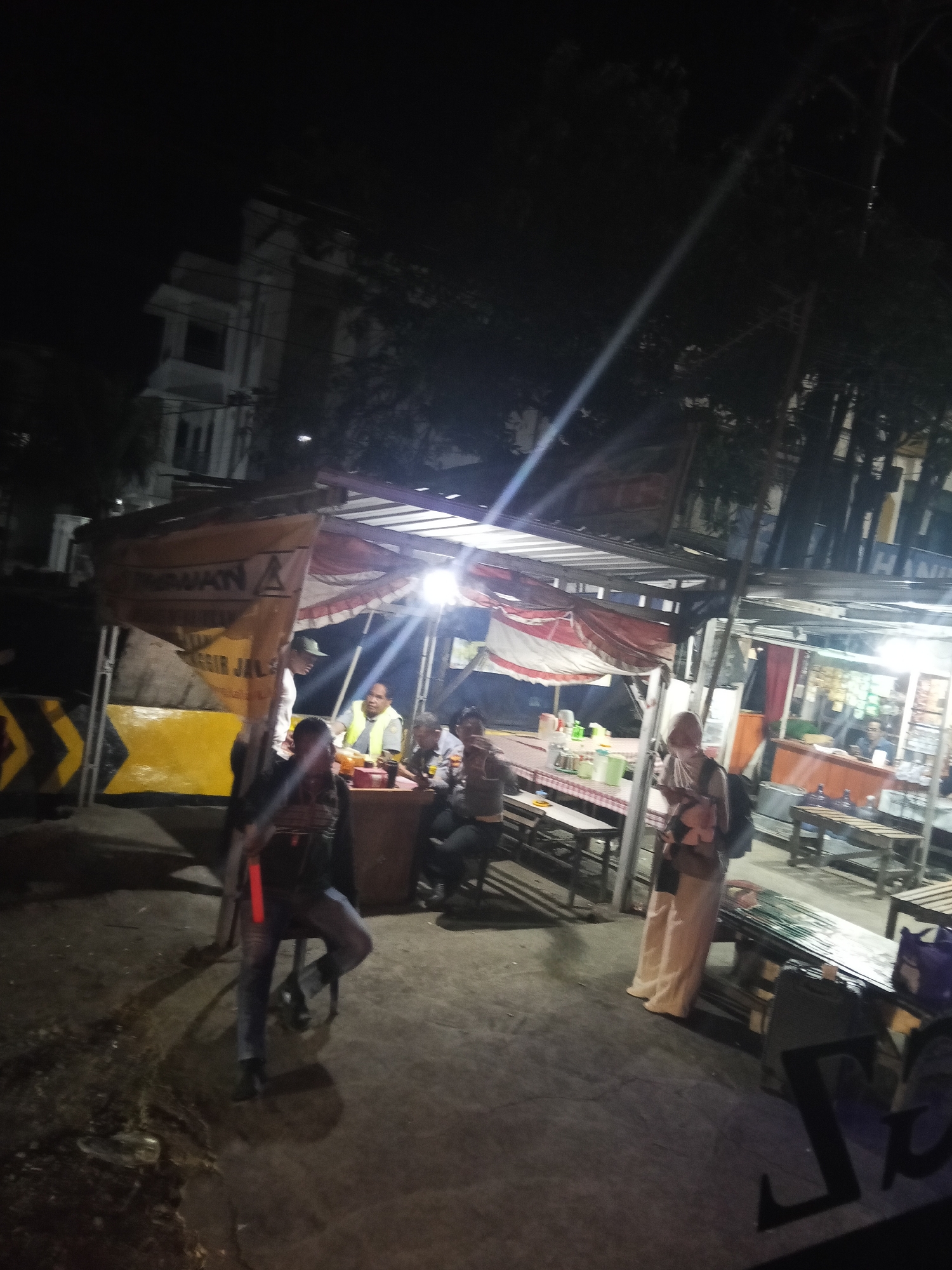 The image captures a street-level view at night, likely in Marumpa, Indonesia, featuring a food stall and its surroundings. In the foreground, a person is seated on a stool, facing away from the camera, with a red object, possibly a stick or baton, held vertically by their side. They are wearing jeans and dark clothing. To the left, a yellow barrier with black chevron markings is visible, suggesting a road or construction area.
Behind the seated individual, a food stall is set up with a covered roof supported by poles. The stall displays various items, and people are interacting within and around it. One person, wearing a yellow vest and a cap, appears to be serving or attending to customers. Other individuals are seated at tables or standing. A banner with Indonesian text, including "LAWANI" and "NGGIR JALAN," is draped over the stall.
Further to the right, another part of the food stall area is illuminated, showing more tables and possibly goods for sale. A person wearing a headscarf and light-colored clothing is standing and looking down at a mobile device. Bottles are visible on tables and on the ground. The overall environment is dimly lit by artificial lights, creating shadows and highlights. The ground appears to be paved or a dirt surface. In the background, a multi-story building is faintly visible through the darkness and foliage.