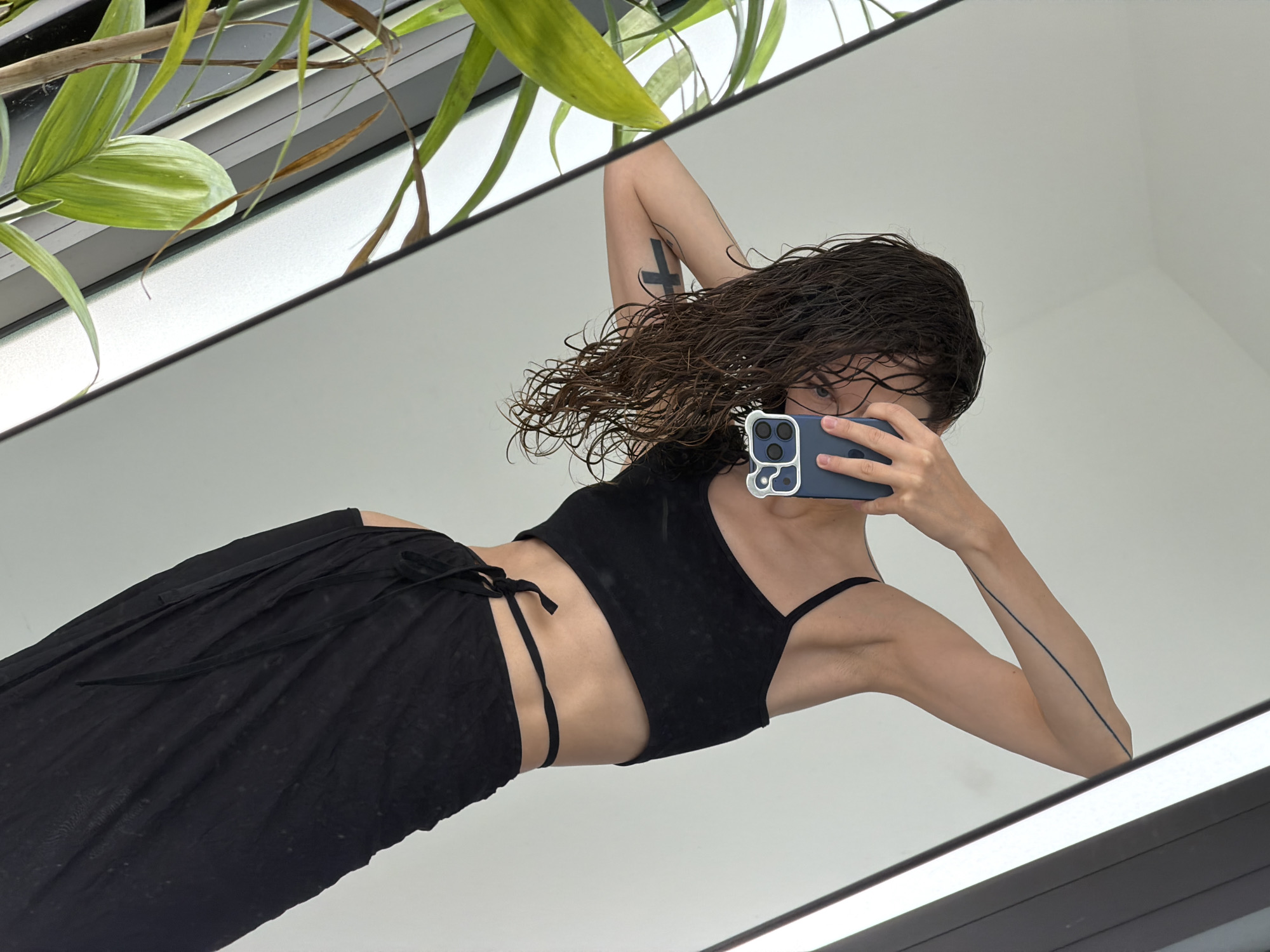 A mirrored self-portrait shows a woman holding a blue smartphone with a cartoon-like sticker on the back casing. She wears a black crop top and a loose, black skirt tied at the waist. Her brown, wet hair is styled loosely. A tattoo of a cross is visible on her right bicep, and another thin, linear tattoo is on her left forearm. Green plants are visible in the top left corner of the frame. The setting appears to be an interior space with white walls and reflective surfaces. There is no verifiable evidence to support the claim of the image originating from Đà Nẵng, Vietnam.