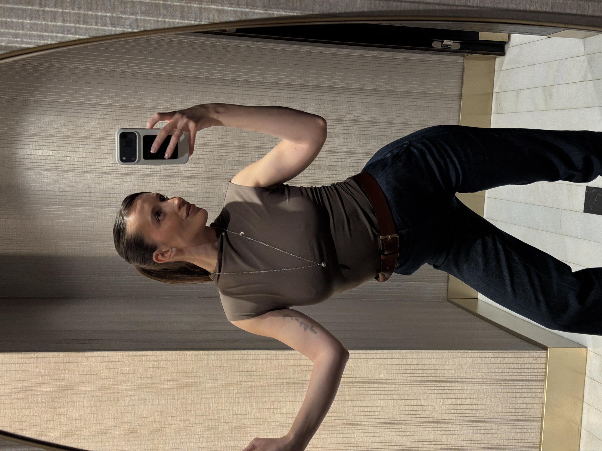 A medium shot depicts a person reflected in a mirror, taking a selfie. The individual has dark hair pulled back into a low ponytail, a brown T-shirt, dark jeans, and a brown belt. They wear a delicate silver necklace and a ring on their left hand. The person is holding a white smartphone with a black camera lens. A tattoo is visible on their left arm.
The reflection is set against a beige, horizontally-striped wall. A light-colored tiled floor with dark accents is visible in the right-hand portion of the reflection, as well as a gold colored border. The setting is indoors. There is no information in the image to indicate this is in Budva, Montenegro.