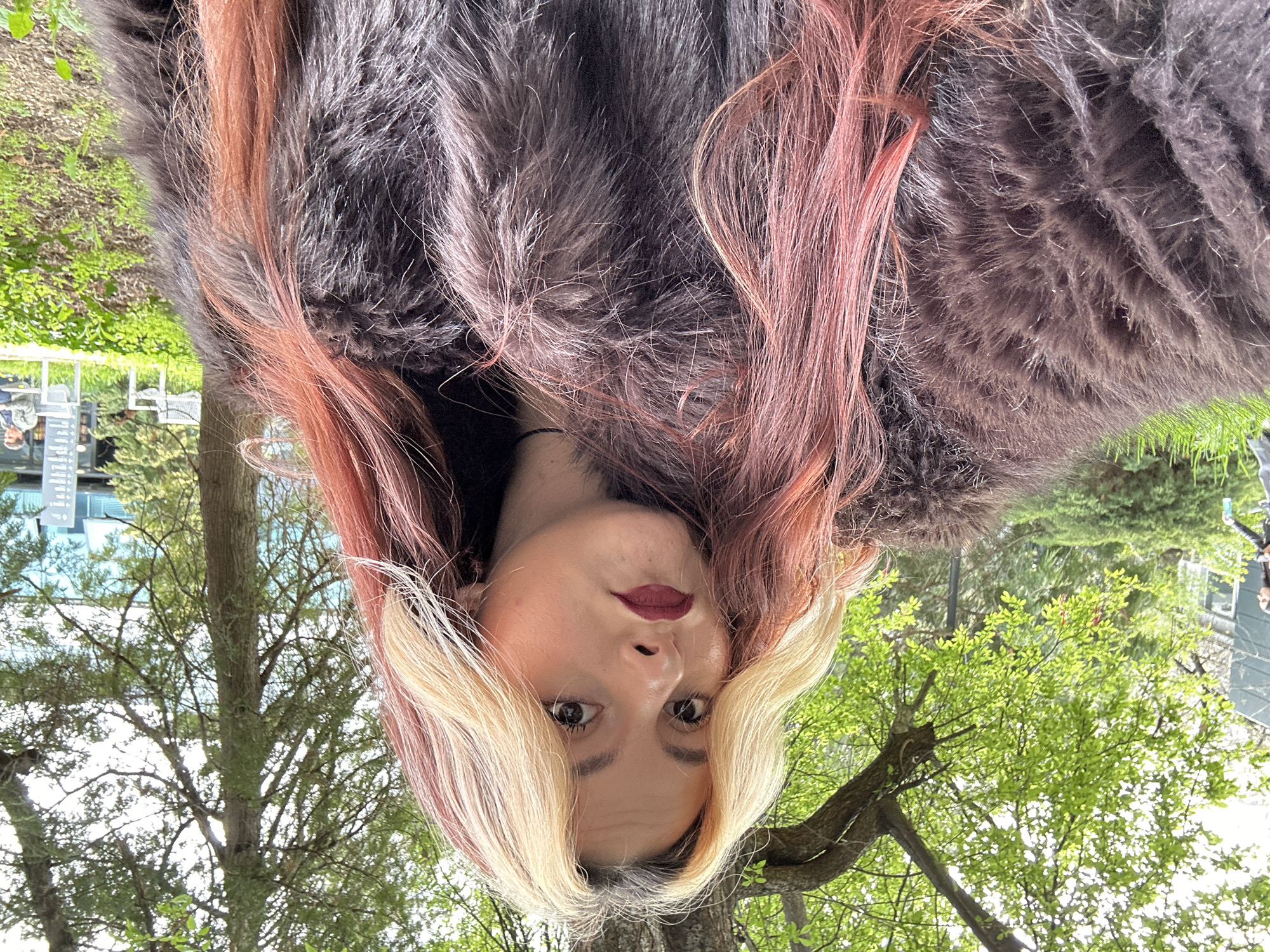 The image presents an upside-down close-up portrait of a person, likely female, framed by their multi-toned hair and a fluffy, dark garment. The individual's hair features prominent dark sections, vibrant pink or reddish highlights, and lighter blonde strands around the face. They look directly at the viewer with a neutral expression, adorned with dark eye makeup and deep red lipstick.
The setting is outdoors, likely a park or urban green space in Sofia, Bulgaria, with the background appearing inverted due to the image's orientation. Lush green trees with abundant foliage and grassy areas fill most of the background, suggesting a spring or summer day. Distant urban elements are faintly visible, including structures that could be buildings or parts of a cafe area with tables and chairs. On one distant building, some unclear repeated symbols or punctuation marks can be seen. The bright, natural lighting indicates daytime. The subject's inverted pose suggests a deliberate artistic or playful choice.