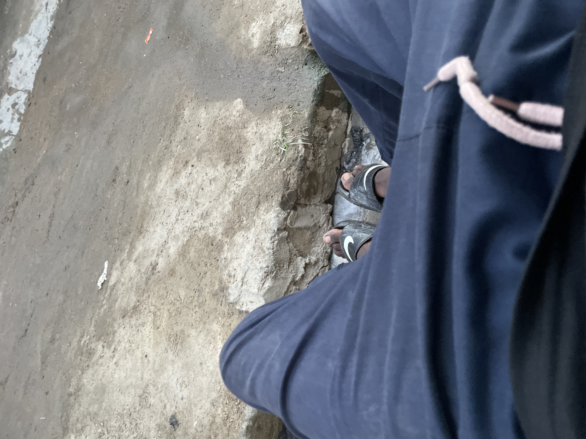 The image captures a downward, high-angle view of a person's lower body and the ground. The subject is wearing dark blue, loose-fitting pants, one leg of which is prominently featured on the right side of the frame. A light-colored drawstring with metal tips is visible on the pants. The person's feet are clad in black slide sandals, one of which clearly displays the white "Nike" swoosh logo.
The setting appears to be an outdoor area with a ground surface composed primarily of dirt and rough, textured concrete or stone. A distinct, uneven curb-like structure runs from the mid-left towards the bottom right of the image, separating a flatter, lighter ground area from a slightly lower, darker patch of soil where a few blades of green grass are sparsely growing. On the lighter ground, a small orange piece of debris and a small white piece of debris are visible. The lighting suggests it is daytime. No specific activity beyond standing or walking is evident. The overall impression is one of an ordinary, utilitarian outdoor environment.
