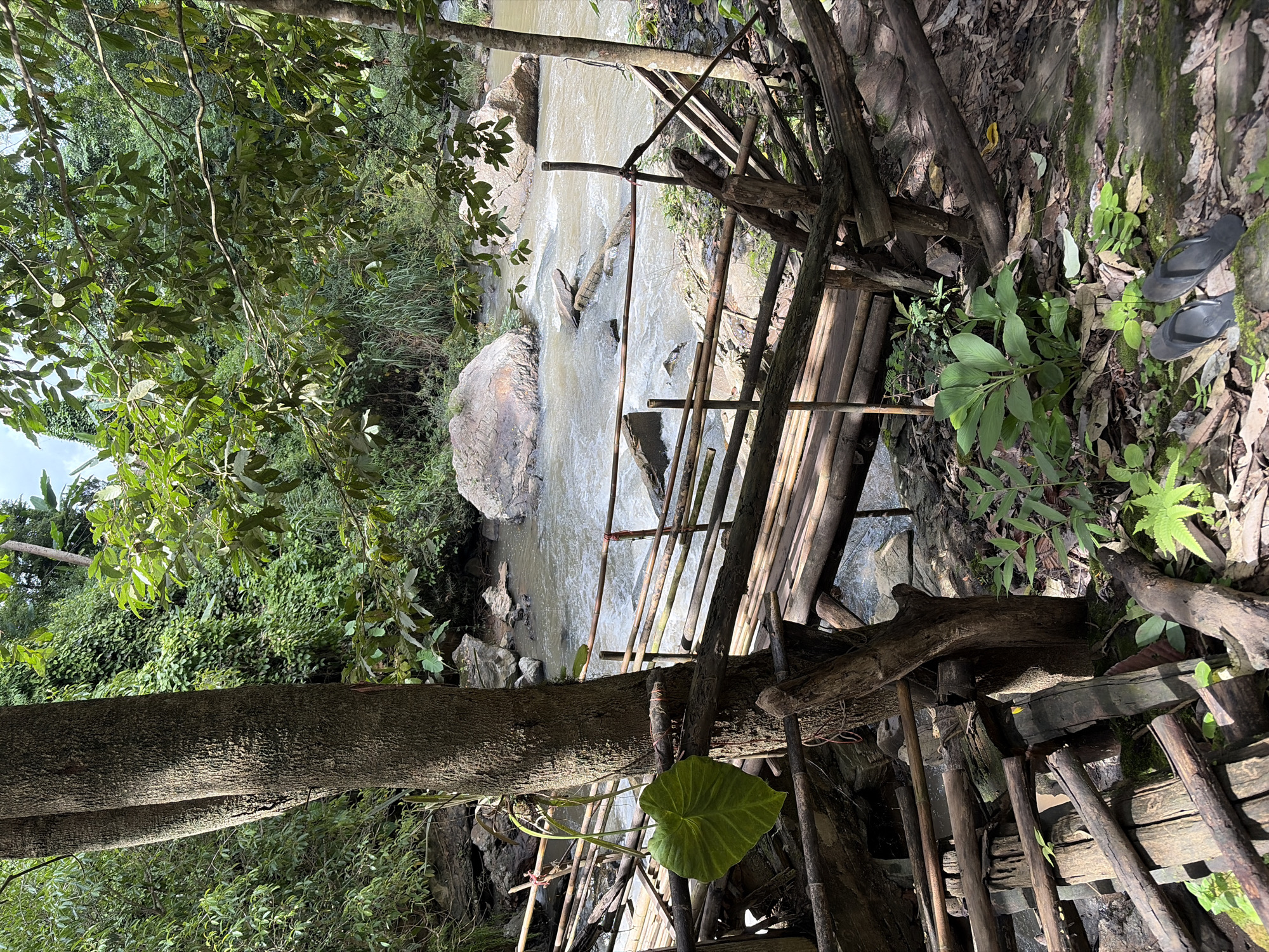 This image depicts a rustic bamboo bridge crossing a shallow, rocky river in the city of Ban Huai Takhrai, Thailand. The bridge, constructed from natural bamboo and logs, appears weathered and somewhat precarious, with a few railings missing or broken. The river flows briskly over smooth, rounded rocks, with larger boulders visible in the water and along the banks. Lush green foliage, including large-leafed plants and ferns, dominates the surrounding environment, with the dense canopy of trees casting shadows on the scene. In the foreground, a pair of dark-colored flip-flops rests on a moss-covered rock. The overall impression is one of a natural, somewhat wild setting, accessible by a rudimentary man-made structure.
