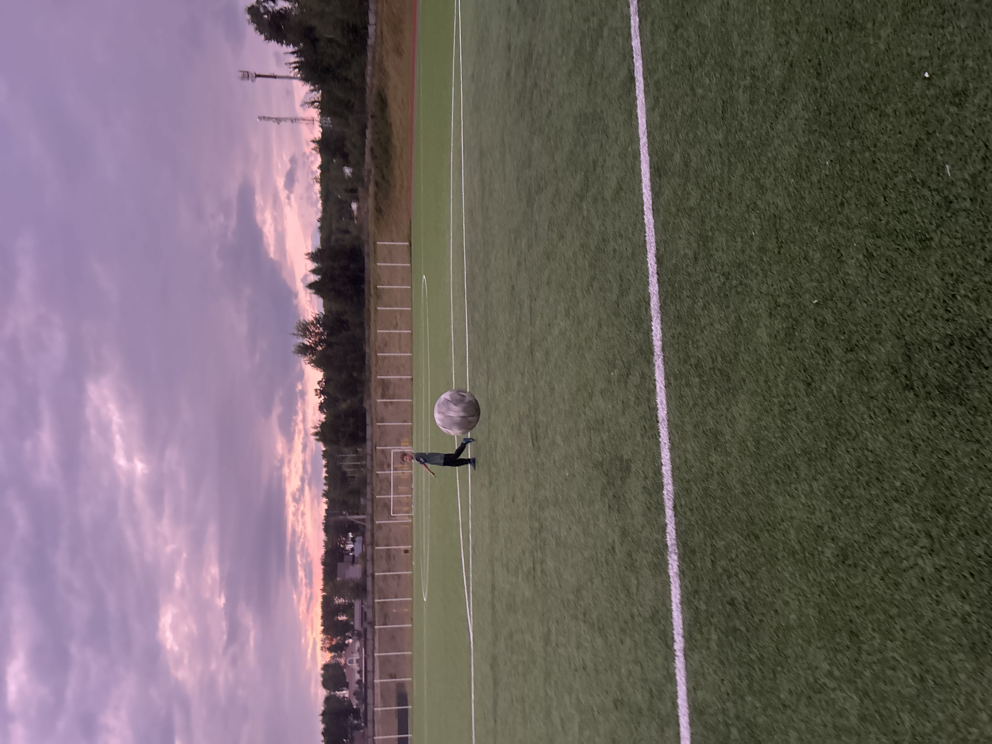 The image depicts a boy on a green, artificial turf sports field, likely a soccer field, given the white markings. The boy, wearing a dark t-shirt and black pants, is running towards the right side of the frame. He is interacting with a large, spherical object that appears to be an inflatable ball, possibly a zorb ball, which is translucent and has a textured surface. In the background, a line of white posts and a fence border the field, beyond which are trees and some distant buildings, including houses and what appear to be communication towers. The sky above is filled with dramatic, purple and pink clouds, suggesting either sunrise or sunset. The overall lighting is dim. The scene is located in Noviy, Russia.
