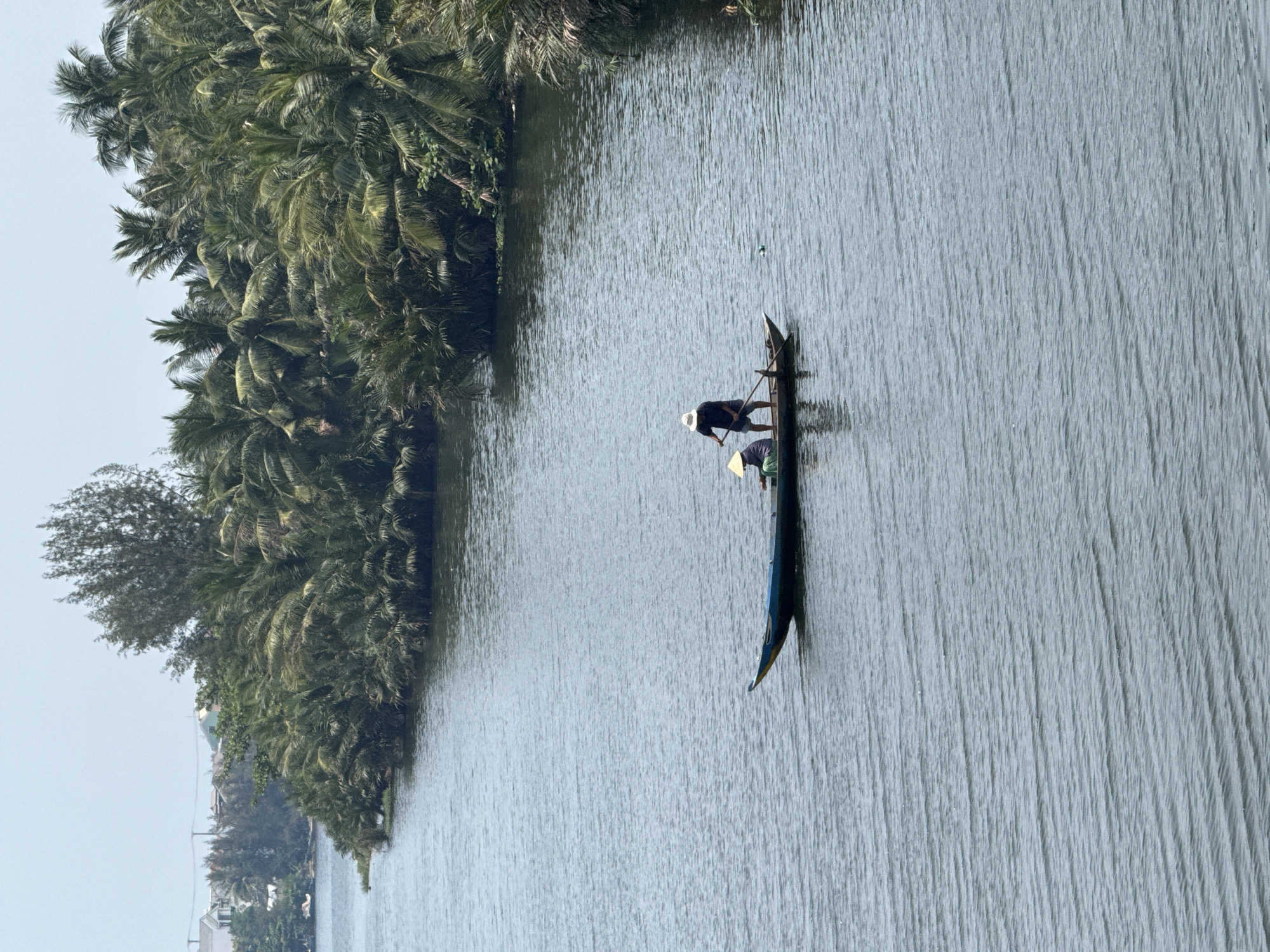 Two people are in a narrow boat on a river. One person is standing and holding a long pole, possibly pushing the boat. The other person is sitting in the boat. The river is lined with trees and vegetation on one side. The river water is gray-blue in color. The scene is likely located in a rural area near Đà Nẵng, Vietnam, as similar waterways and boats are common in the region.