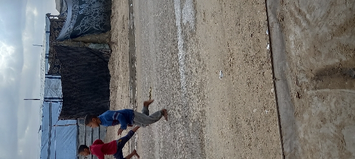 The image depicts an outdoor scene in Khan Yunis, Palestinian Territory, under an overcast sky. In the foreground, two young males, appearing to be children, are running from left to right across a dirt and sand surface containing patches of moisture. The child on the left wears a red long-sleeved shirt and dark pants, running barefoot with his left leg forward. The child on the right, slightly behind, wears a blue long-sleeved shirt with light stripes and grey pants, also barefoot, with his right leg forward. Both children are in mid-stride, looking forward.
The background features several makeshift structures consistent with a temporary settlement or camp. These structures are constructed from various materials, including light blue tarpaulins and dark patterned fabrics. One prominent dark fabric structure on the right has a partially visible figure behind it. The ground transitions from a rough, dirty surface in the foreground, marked by a concrete curb or low barrier, to a similar sandy terrain where the structures are erected.
