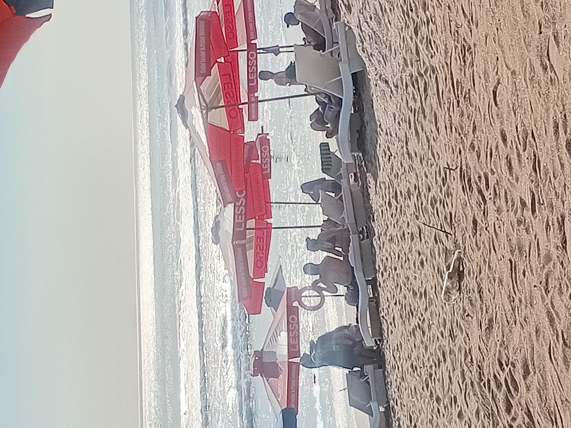 This image captures a beach scene in Kuta, Indonesia, featuring several red beach umbrellas with the word "LESSO" visible on them. Underneath one of the umbrellas, a group of people are seated on lounge chairs. Another person is standing near the water, and a circular flotation device is held by someone. The ocean stretches out in the background with gentle waves, and the sand in the foreground is textured with ripples. A discarded white object, possibly plastic, lies on the sand.