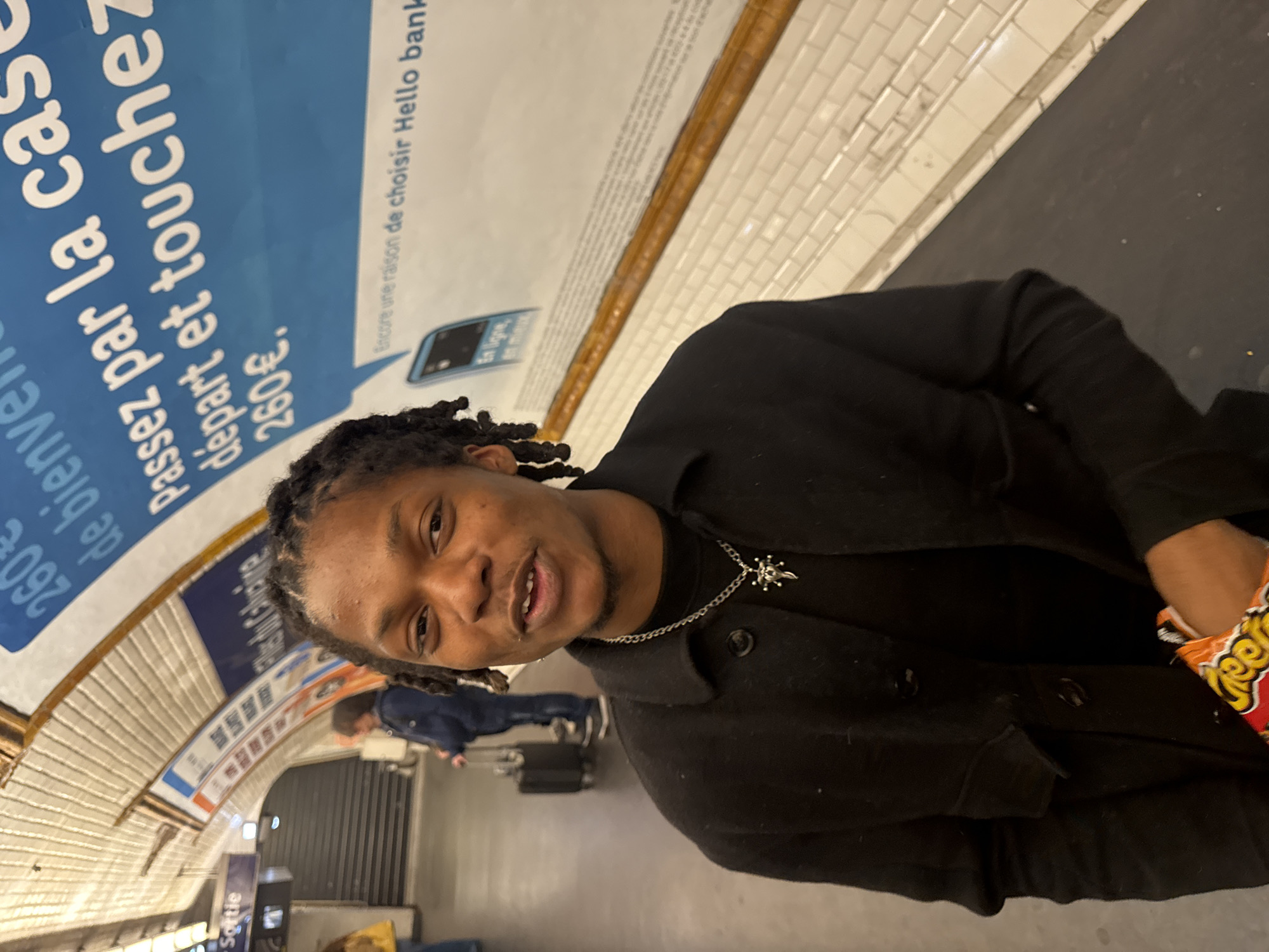 The image appears to be taken in a subway station. A man wearing a black jacket and a silver chain necklace is prominently featured. The man is holding a bag of Cheetos. In the background, there are people with luggage, suggesting the location is a transit hub. A blue advertisement with French text, "Passez par la case départ et touchez 260€", is visible on the wall, likely an advertisement for a bank, "Hello bank." The wall is lined with white tiles.