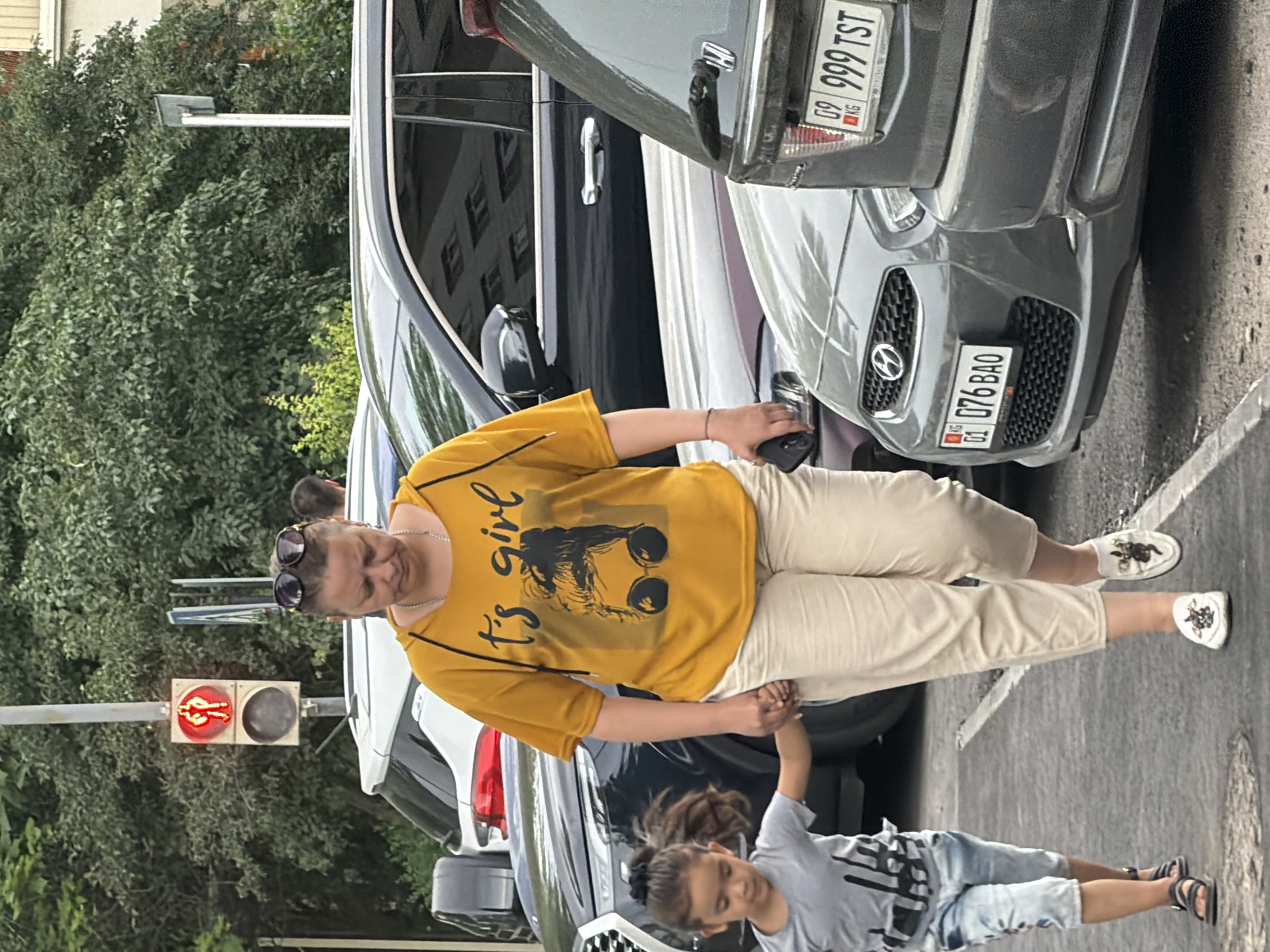 A woman in a yellow hooded shirt with the graphic "it's girl" and a picture of a woman's face with sunglasses is walking on a paved surface, holding hands with a young child. The child is wearing a grey t-shirt and denim shorts. The woman is wearing light-colored pants and white loafers with a black and gold embroidered design. They are walking past a row of parked cars, including a dark-colored SUV and a grey sedan. In the background, a pedestrian traffic light shows a red walking figure. The environment appears to be an urban setting with trees and buildings visible.