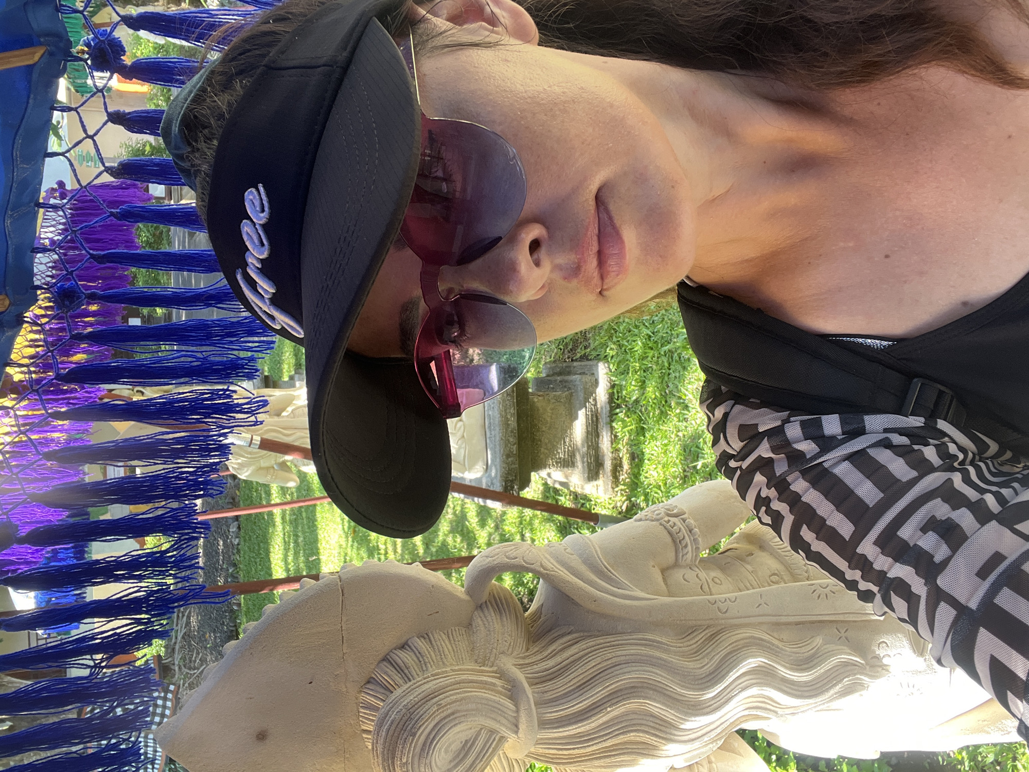 A woman wearing a black visor with "free" embroidered on it and pink-tinted sunglasses takes a selfie outdoors. She is wearing a black top with a sheer, patterned overlay. To her left, a partially visible light-colored stone statue of a person with ornate hair is positioned. The background features lush green grass, a stone pedestal, and a canopy with hanging blue and purple tassels. The scene appears to be set in an outdoor area, potentially a temple or cultural site in Kuta Selatan, Indonesia.