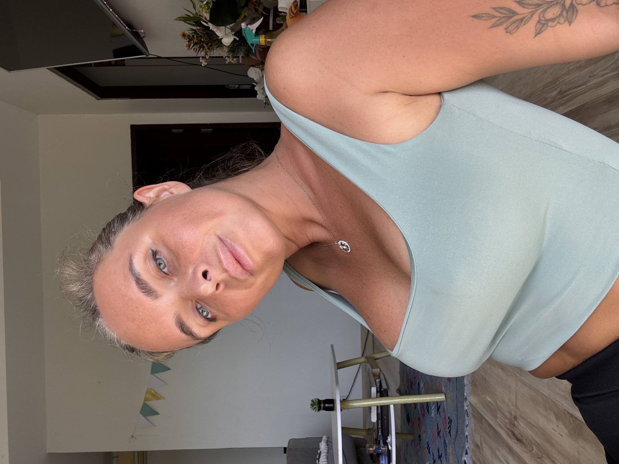 A woman is photographed from the chest up, in a selfie taken indoors. She is wearing a light blue tank top and a silver necklace with a small pendant. Her hair is pulled back, and she has a light tan. The background shows a living room with a couch, a coffee table with a small plant, and a television mounted on the wall. There are also some decorative flags hanging on the wall. The woman appears to be in Baan Chaweng, Thailand, based on contextual information.