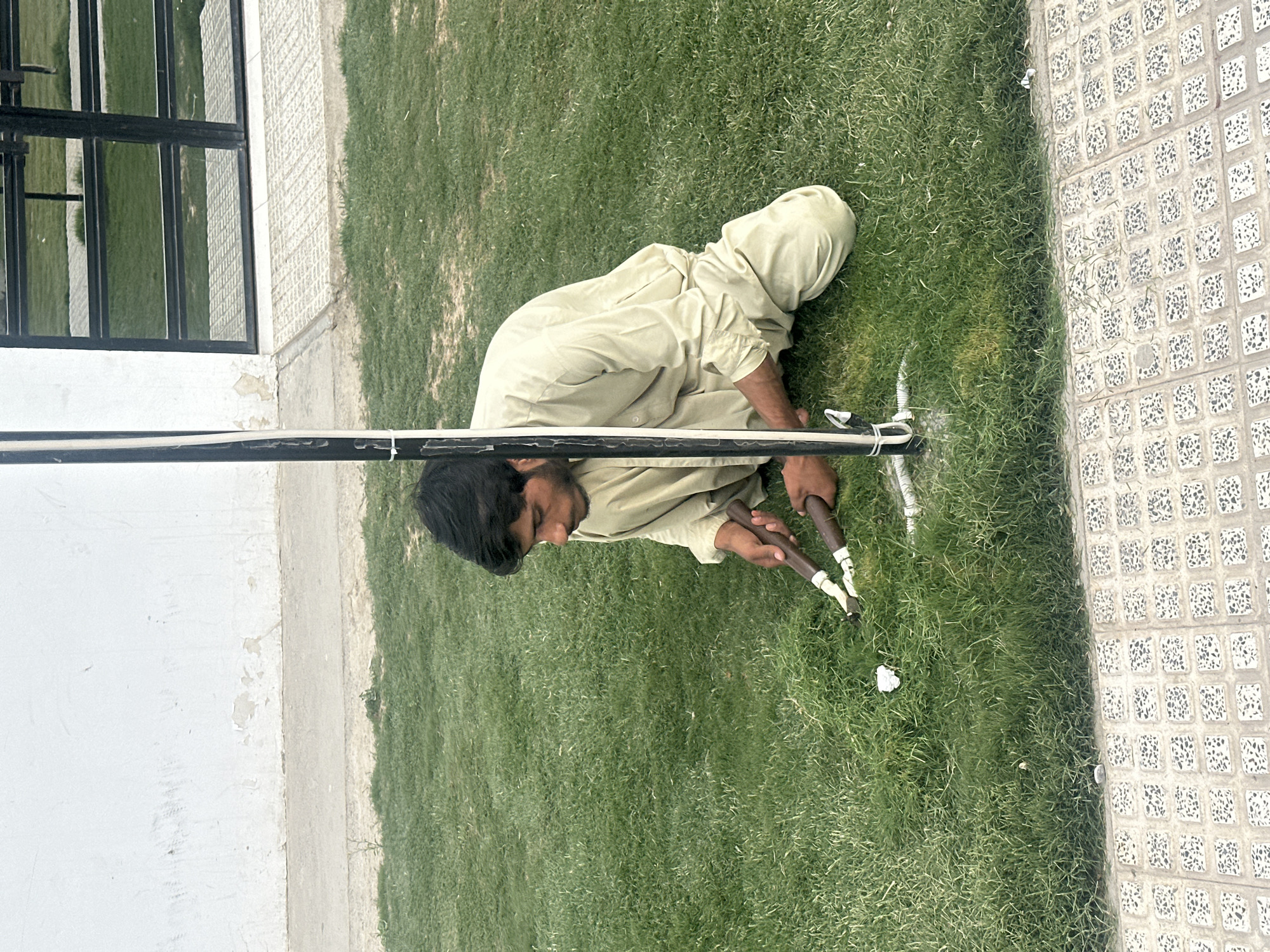 A Pakistani male kneels on green grass between a white-painted building with black metal window frames and a patterned tiled sidewalk in Quetta City Tehsil, Pakistan. He wears a light-colored traditional shalwar kameez. He uses a pruning tool to trim the grass. A long, thin, dark pole, secured with white cable ties, is visible in the foreground.