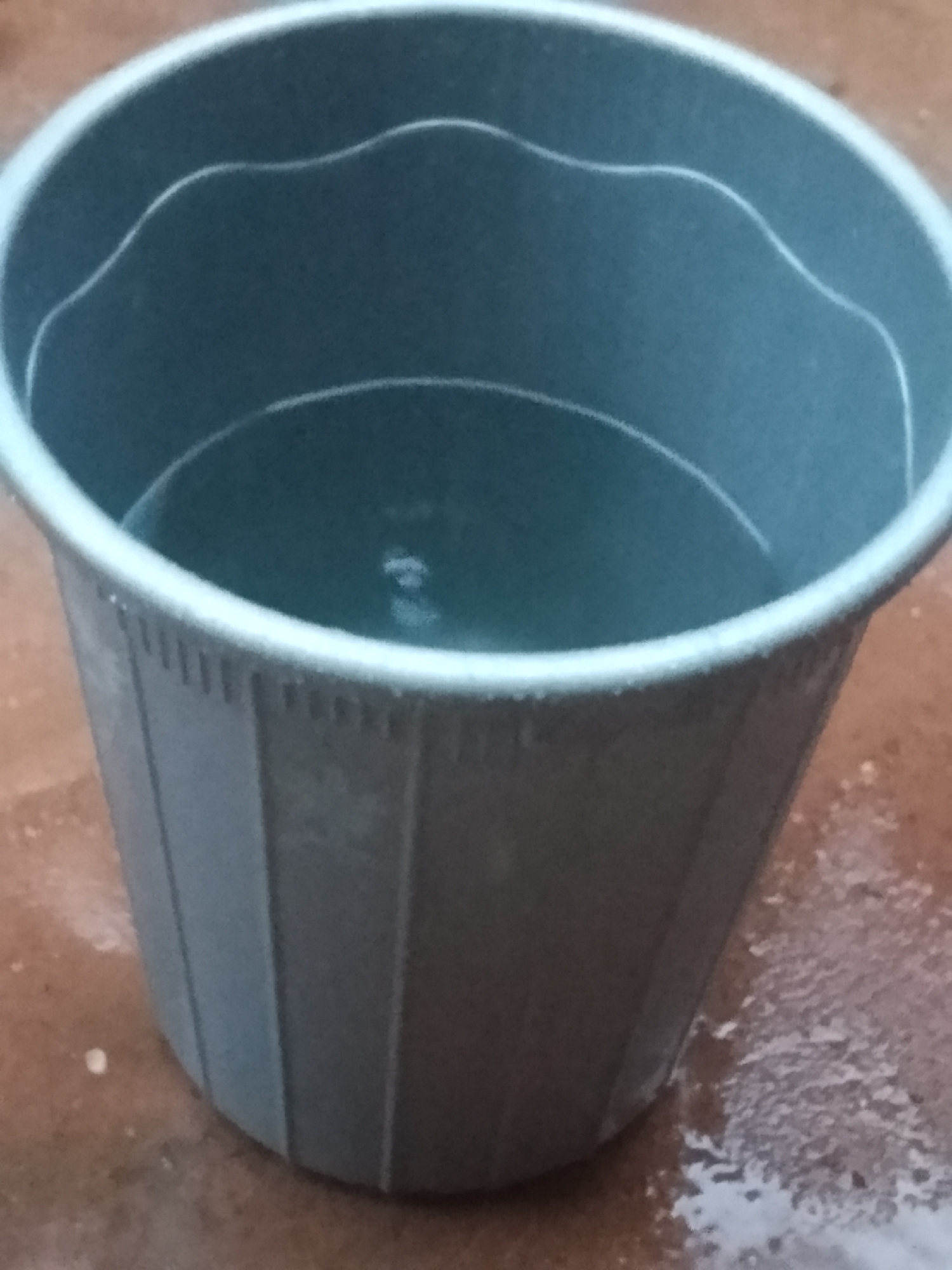 A grey plastic bucket is filled with a small amount of clear water. The bucket has vertical ridges along its sides and a wavy rim. The bucket is placed on a damp, reddish-brown surface that appears to be concrete or dirt. There are small droplets of water scattered on the surface around the bucket. The image is taken from a slightly elevated, angled perspective, looking down into the bucket. The lighting is neutral, and there are no people or other objects visible in the scene.