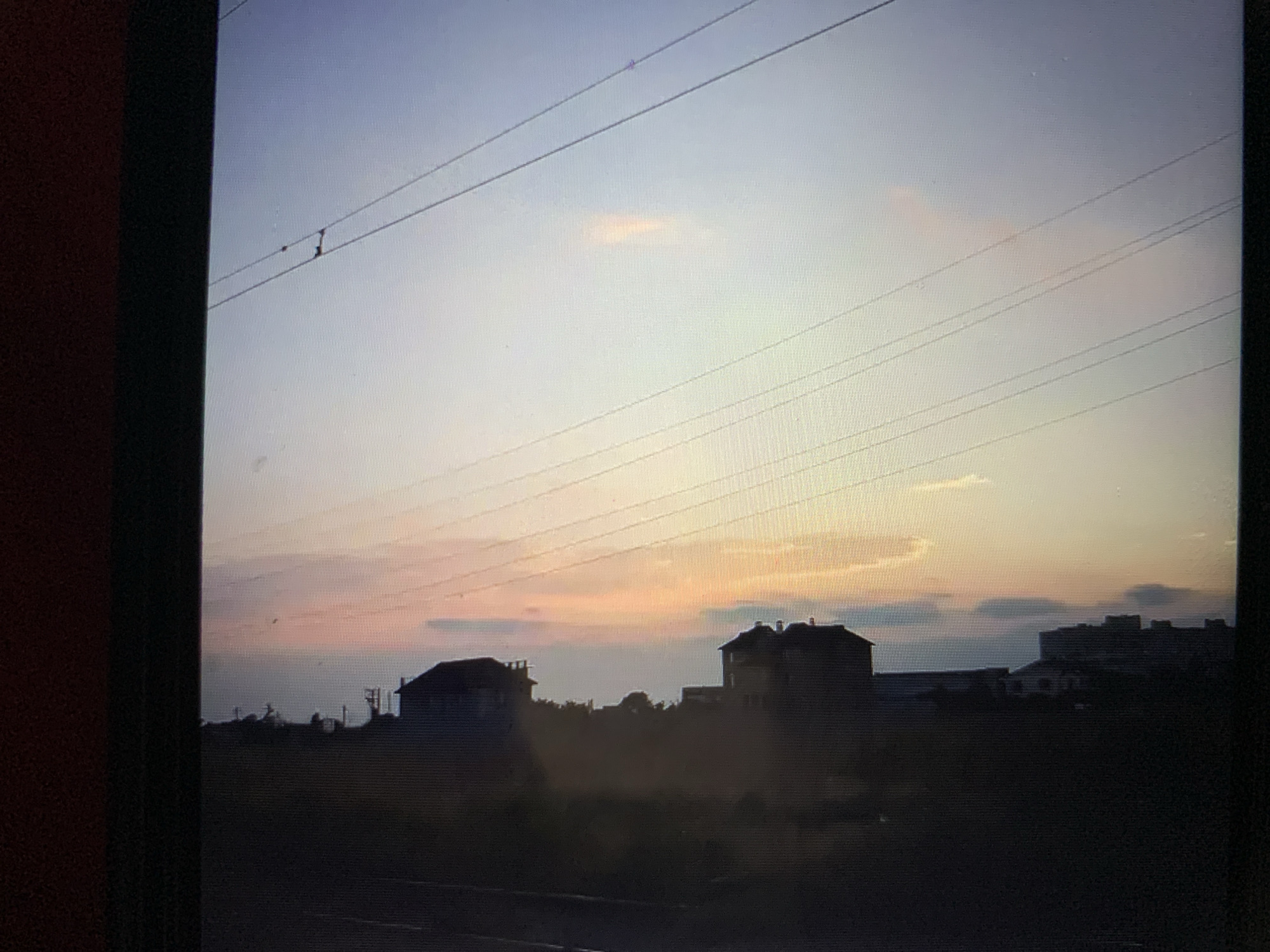 The image displays a silhouetted cityscape with buildings against a twilight sky in Lymanka, Ukraine. Multiple utility wires crisscross the frame, stretching across the upper right portion of the image. The sky is a gradient of colors, with soft pinks and oranges blending into a pale blue. Some of the buildings have visible architectural details, suggesting they are residential or commercial structures. The overall impression is a quiet, urban scene at dusk, likely captured from a distance.
