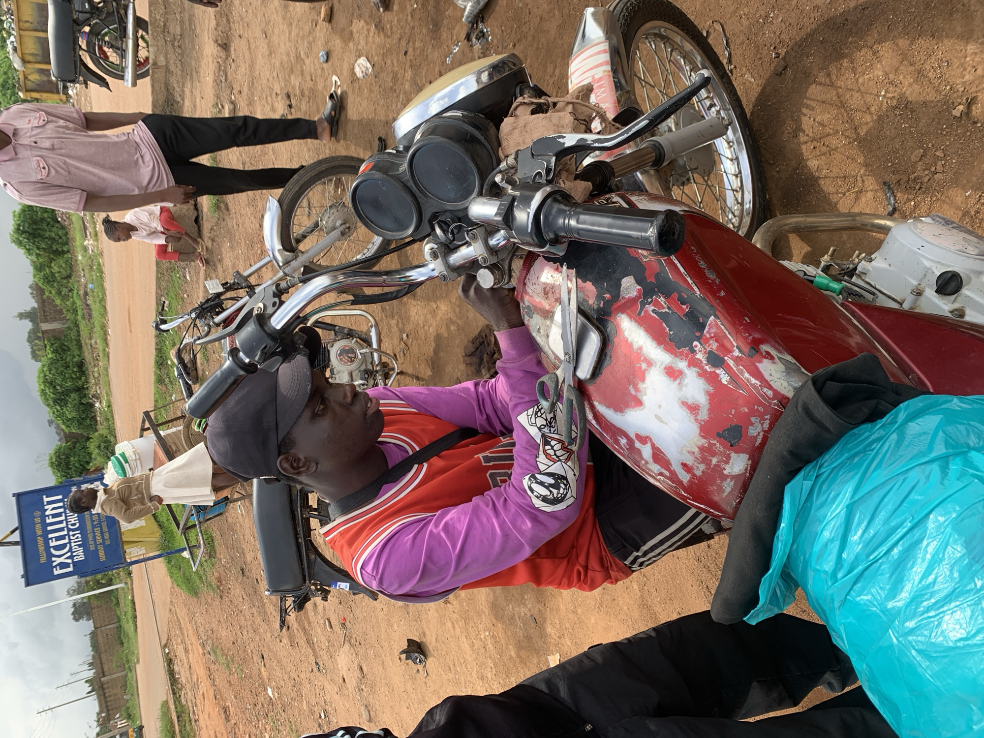 A young Black male wearing a red and purple basketball jersey and black athletic shorts with white stripes is working on a red motorcycle. He is holding scissors and appears to be cutting something on the motorcycle. The motorcycle has a worn, scratched paint job with red and white accents. Behind the motorcycle, a blue sign with white lettering reads "EXCELLENT BAPTIST CHURCH." A young Black woman in a tan jacket and white skirt stands near the sign, looking away from the camera. Another young Black child in a light pink shirt and red pants sits on a concrete barrier in the background. A man in a pink polo shirt and black pants stands to the right of the child. The ground is dirt and gravel, suggesting an outdoor, possibly rural or roadside setting. In the far background, a yellow dumpster and other motorcycles are visible. A bright blue plastic bag is placed on the motorcycle's seat. The sky is overcast. The scene appears to be in Barakin Lerawi, Nigeria.