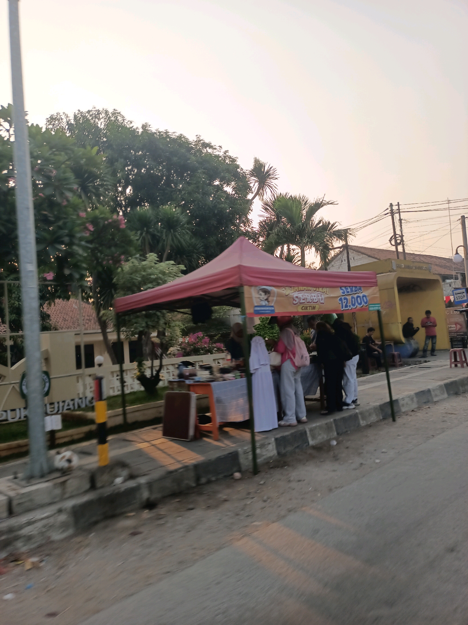 The image depicts a street scene in Karawang, Indonesia, likely in the late afternoon given the warm, soft light. A red canopy tent is set up on the sidewalk, serving as a stall. Underneath the tent, several people are gathered, appearing to be customers or vendors. One person is wearing a pink hijab and a backpack, and another is in a white hijab. The stall has a sign that reads "SERBU 12.000" and an image of a cartoon girl. To the right of the stall, there is a yellow bus stop shelter. In the background, trees and buildings are visible, along with power lines. On the sidewalk near the stall, there are a couple of cats. The street in the foreground is paved, and there is a concrete curb with some debris on it. A gray pole with a yellow and black striped marking is also visible on the left side of the frame.