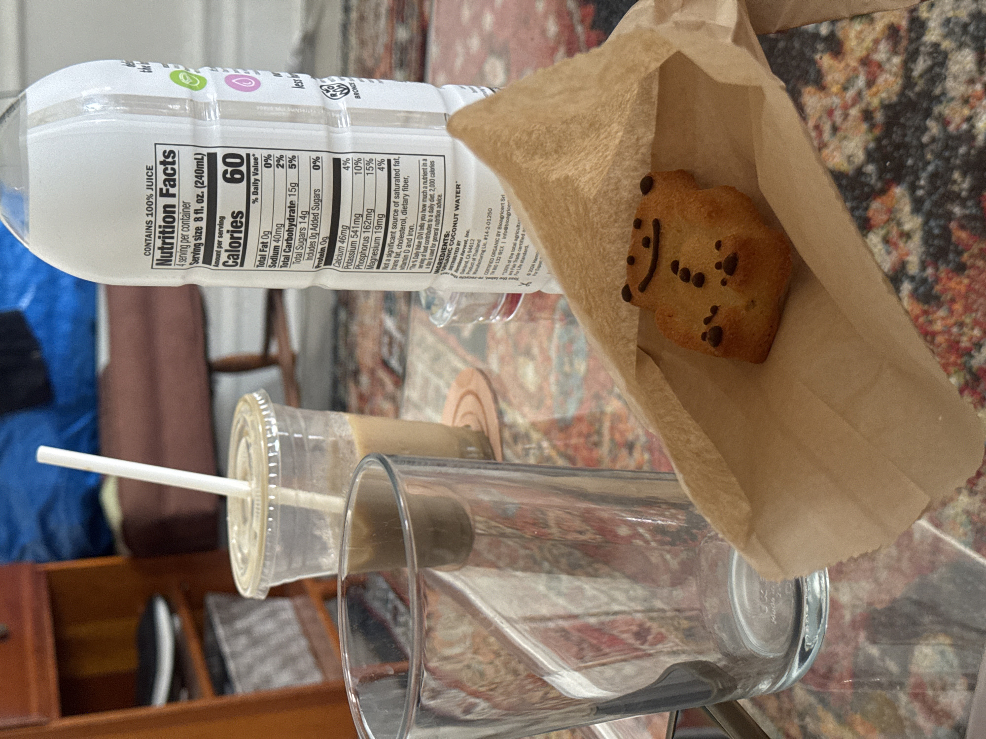 The image shows a tabletop scene, likely indoors, with several food and beverage items. A bottle of "Harmeless Harvest" organic coconut water with visible nutrition facts and ingredient details is partially visible. Next to it, a brown paper bag holds a cookie decorated with chocolate chips to resemble a smiley face. A plastic cup with a straw and a clear empty glass are also on the table. The surface appears to be a patterned rug or table covering, possibly located within a residential or commercial space within the city of New York, United States of America.