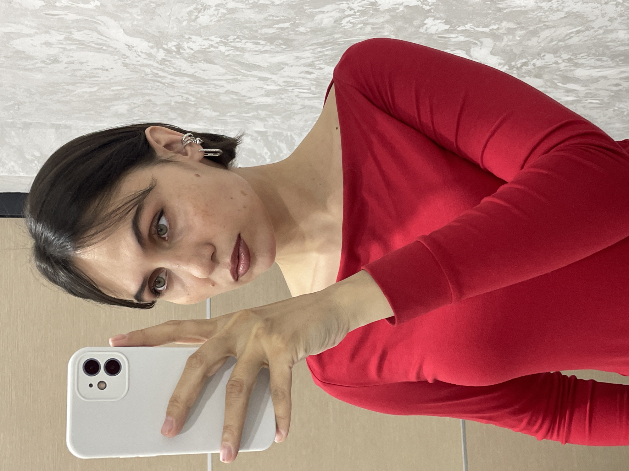 A woman is taking a selfie in what appears to be a bathroom. She is wearing a red long-sleeved shirt and has dark hair. She is holding a white smartphone in her left hand. Her right ear has multiple piercings and earrings. The background shows tiled walls, suggesting a bathroom setting. There's no evidence to confirm the location is Izhevsk, Russia.