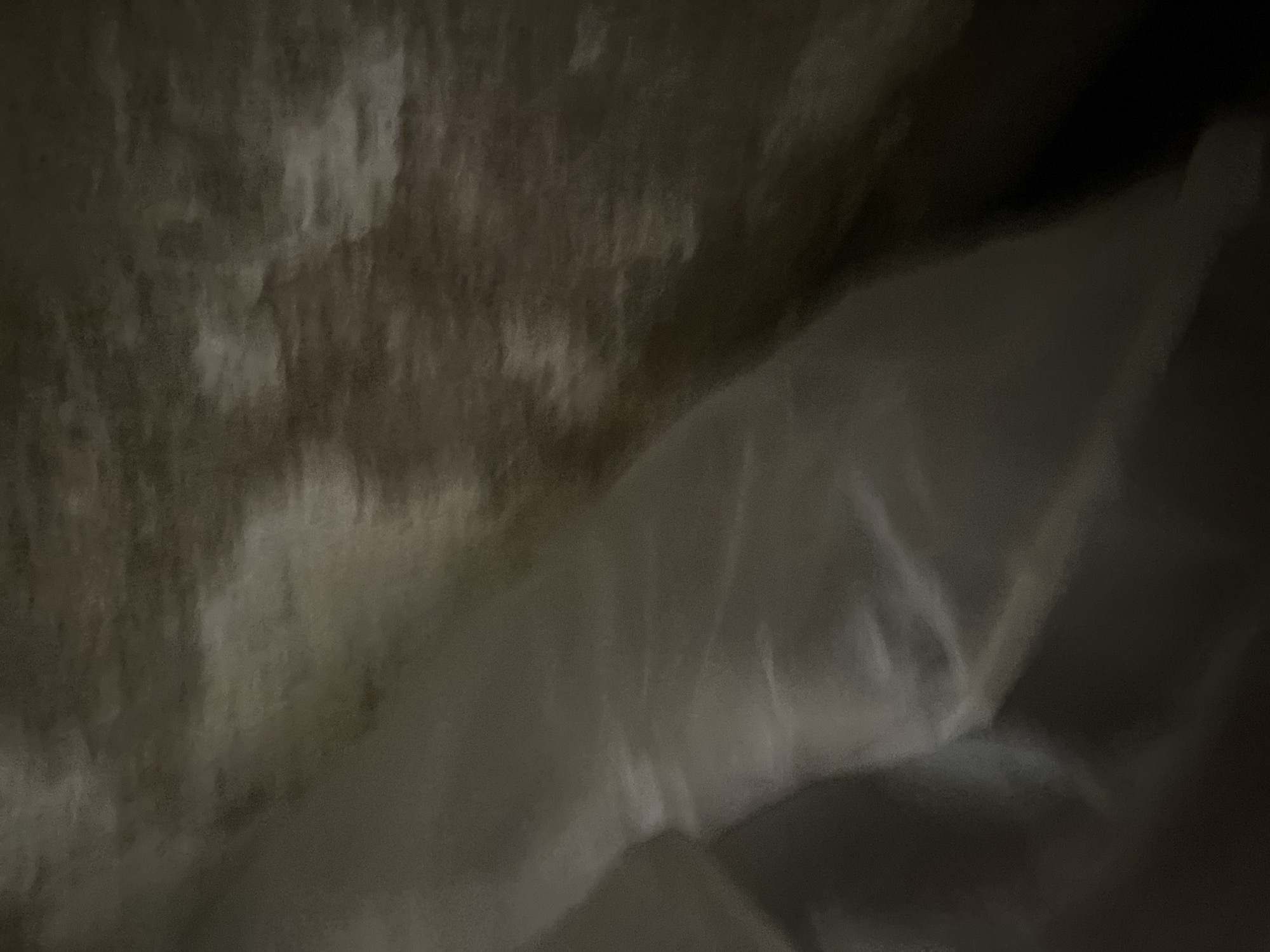 The image is a close-up, abstract shot of textured fabric, likely a blanket or comforter. The colors are muted, ranging from light beige and off-white to dark brown and black, with a fuzzy, almost grainy texture throughout. The fabric appears to be folded or bunched, creating soft shadows and highlights that give it a sense of depth and dimension. There are no discernible objects or people within the frame. The lighting is dim, casting a soft, diffused glow across the surface. The image is entirely devoid of any context relating to Jauro Yinu, Nigeria, or any other specific location.