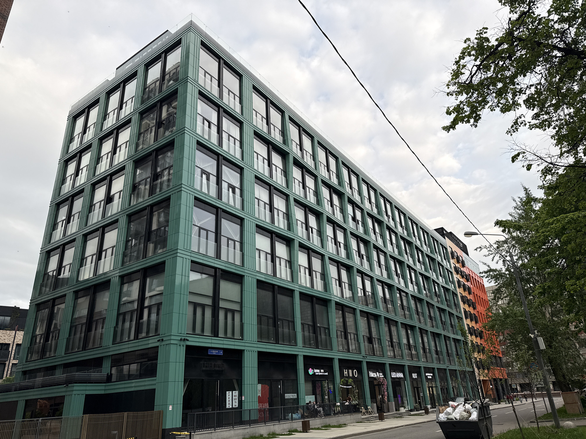 The image shows a street scene in Moscow, Russia. A large, modern building with a distinctive green façade dominates the left side of the frame. This building has multiple stories, with a grid-like pattern of windows. At street level, there are storefronts with signage, including one with the letters "HIIO". To the right of the green building is another building with an orange façade. Trees with green foliage line the street. A utility pole and power lines are visible above. In the foreground, there is a filled trash container on the side of the road. The sky is overcast with cloudy conditions.