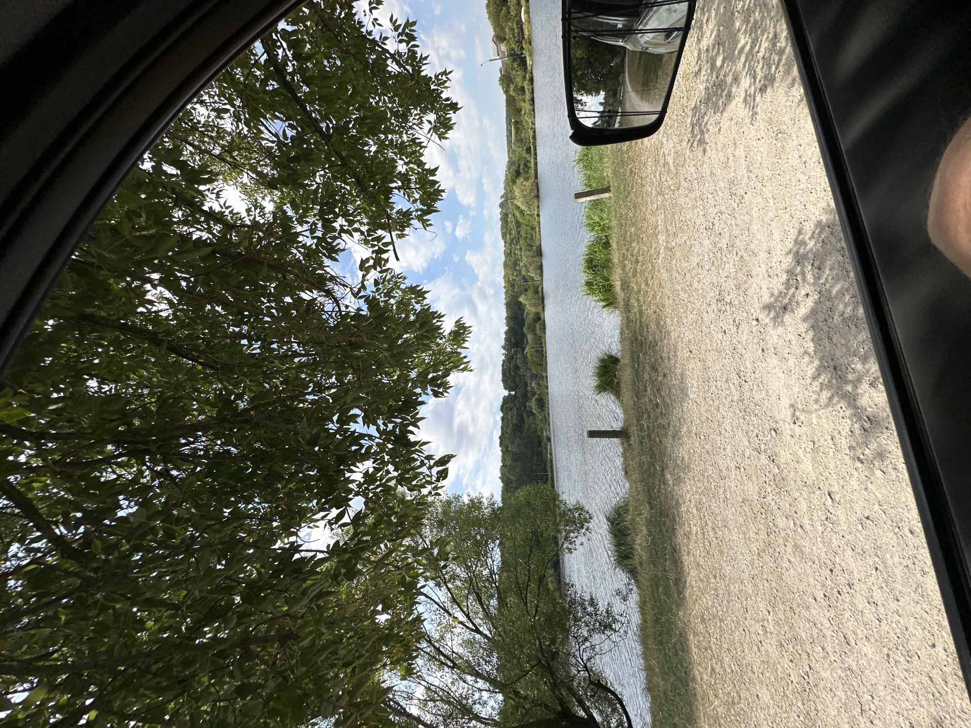 The image captures a view from inside a vehicle looking out towards a body of water. A tree with lush green foliage overhangs the scene, its branches and leaves partially obscuring the sky. The water, appearing calm with ripples, is bordered by a grassy bank and a gravelly path. In the distance, a tree-covered hillside rises, dotted with a few buildings. A side mirror of the vehicle is visible in the lower right corner, reflecting the surrounding landscape and the vehicle's interior. The sky is blue with scattered white clouds. The scene appears to be located near a lake or reservoir in or around Makiv, Ukraine, suggesting a natural or semi-natural environment.