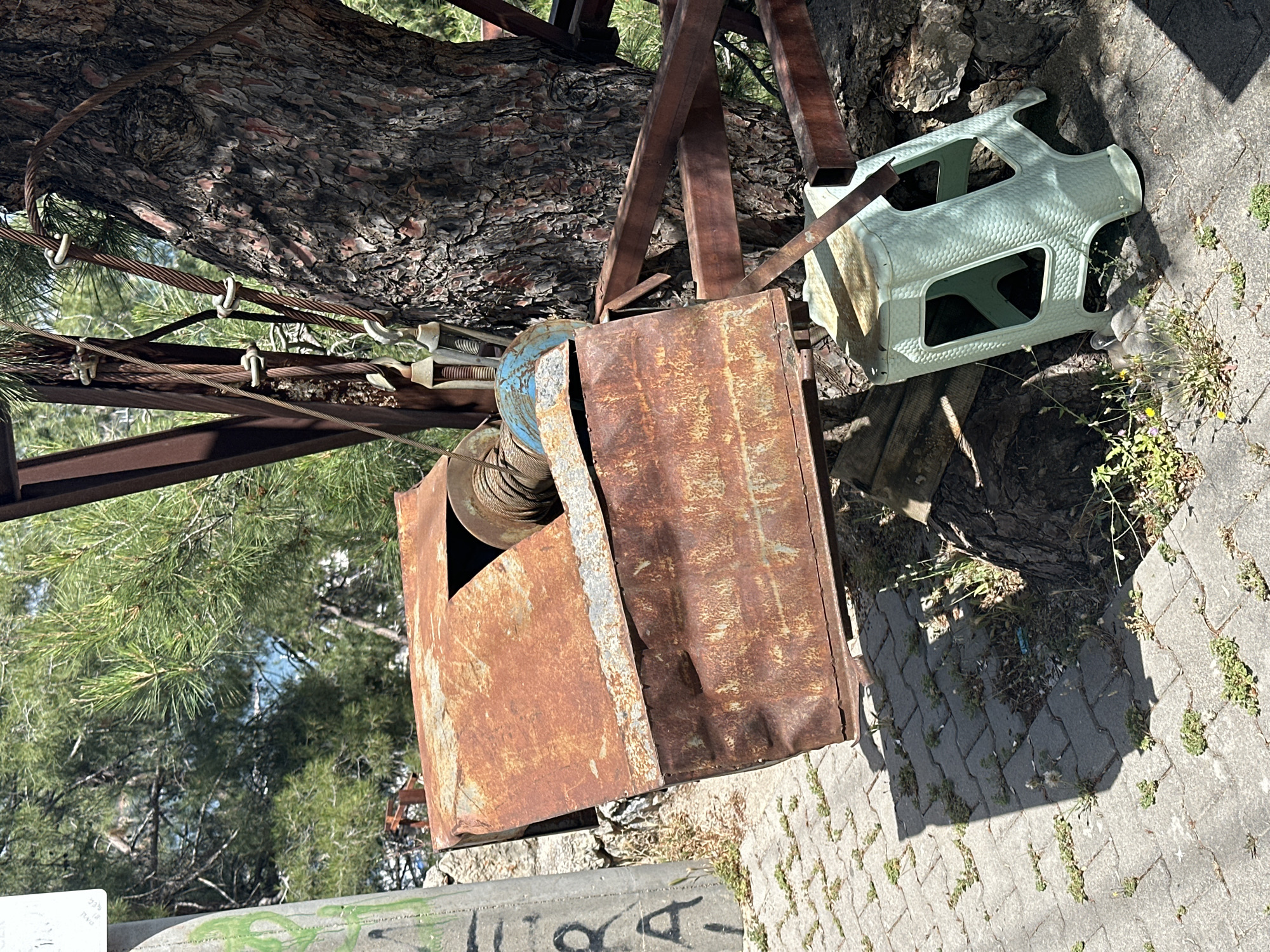 The image shows an outdoor scene in Fethiye, Turkey. A rusty metal box sits near a tree, and a light green plastic chair is placed nearby. The environment includes a tree with thick bark, metal support structures, and paved ground with scattered weeds and small plants. There are no people visible in the image. The overall impression is one of disuse or storage in an outdoor space.