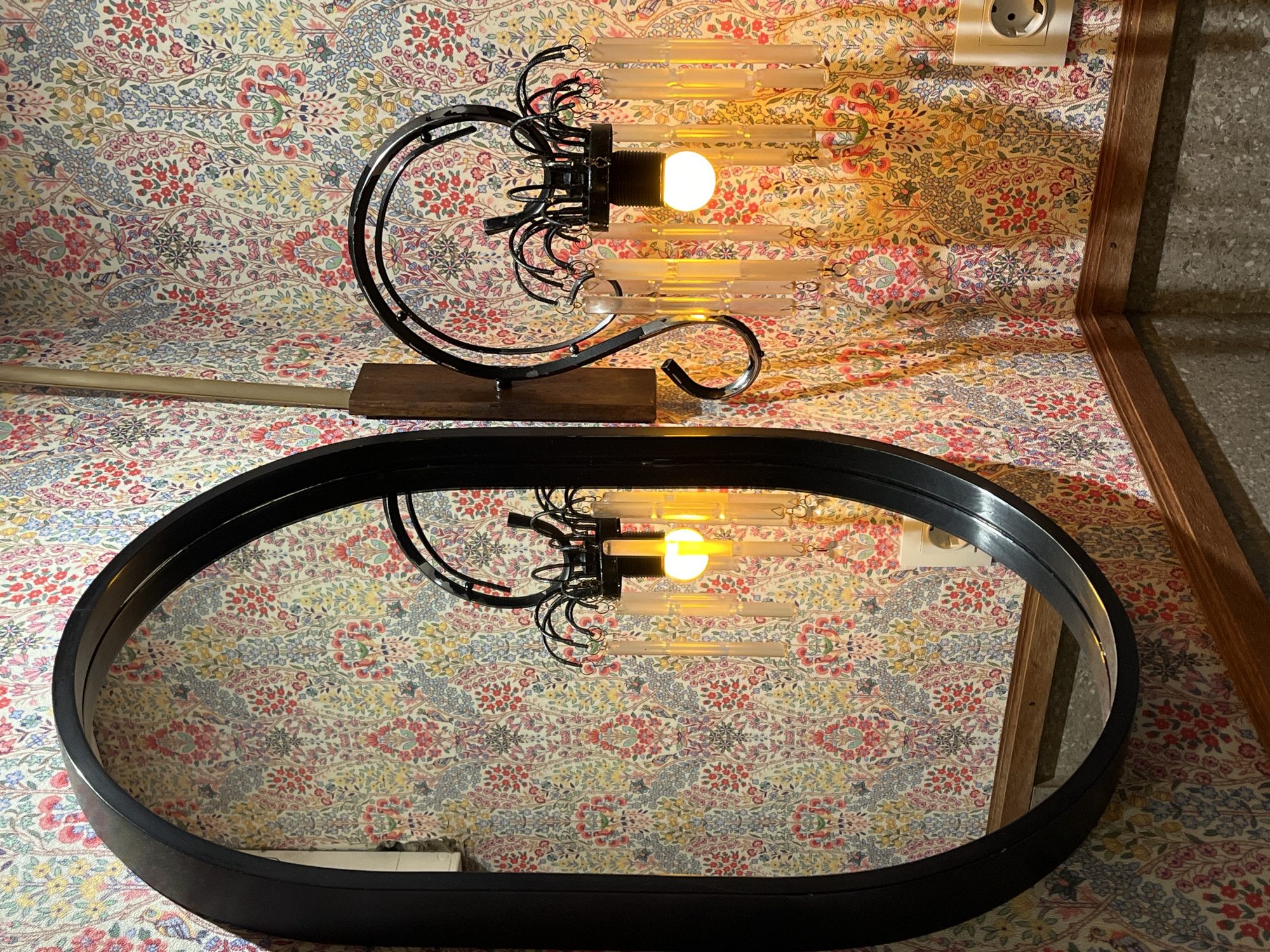 A table lamp is positioned atop a table covered in floral-patterned fabric. Below the lamp, an oval mirror reflects the lamp and the floral fabric. A wall socket is visible in the upper right corner of the image. The location, Denpasar, Indonesia, is not ascertainable from the image.